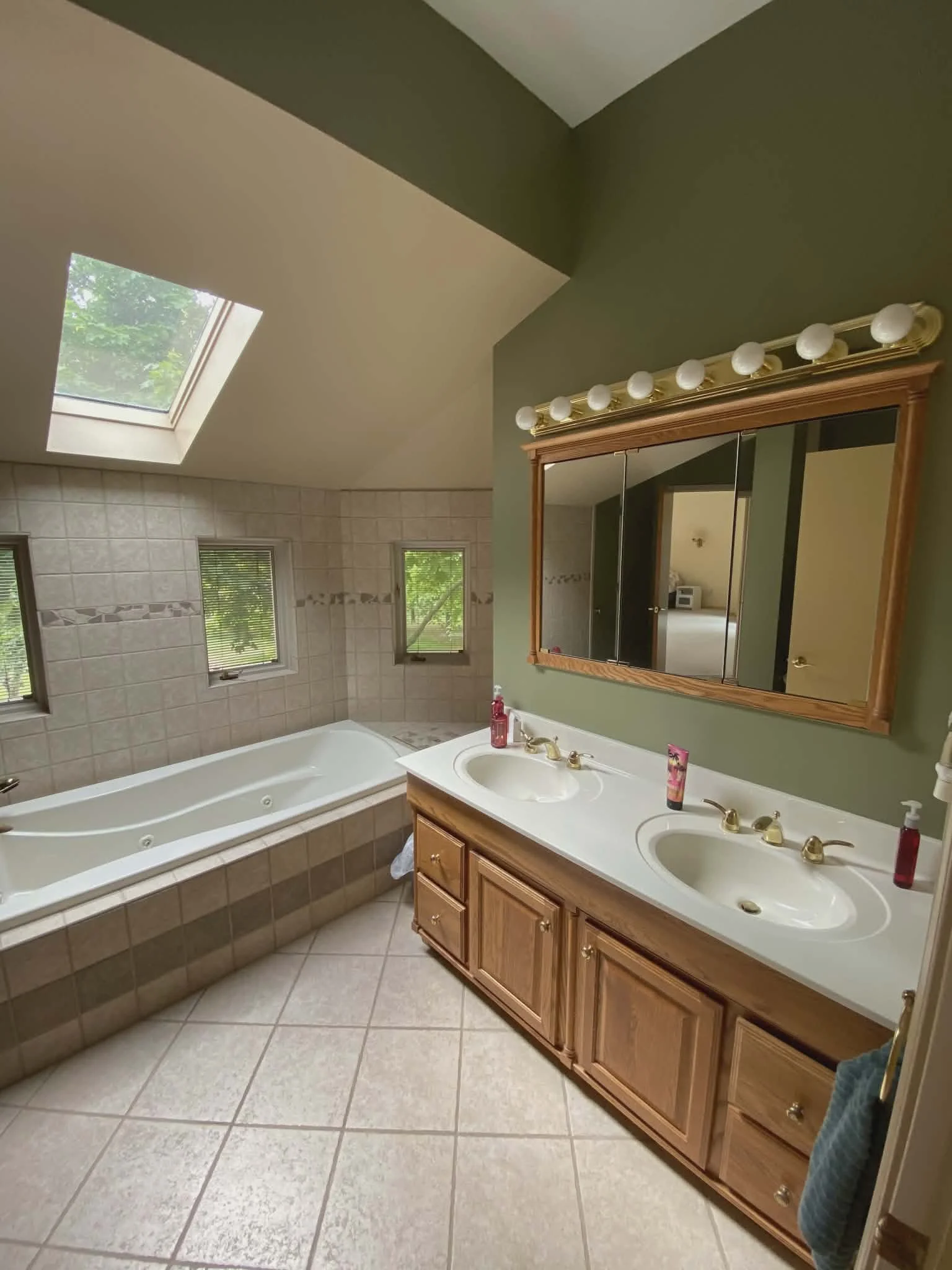 Bathroom with dual sinks, wooden cabinet, mirror, and vanity lights. Bathtub with three windows and skylight overhead.