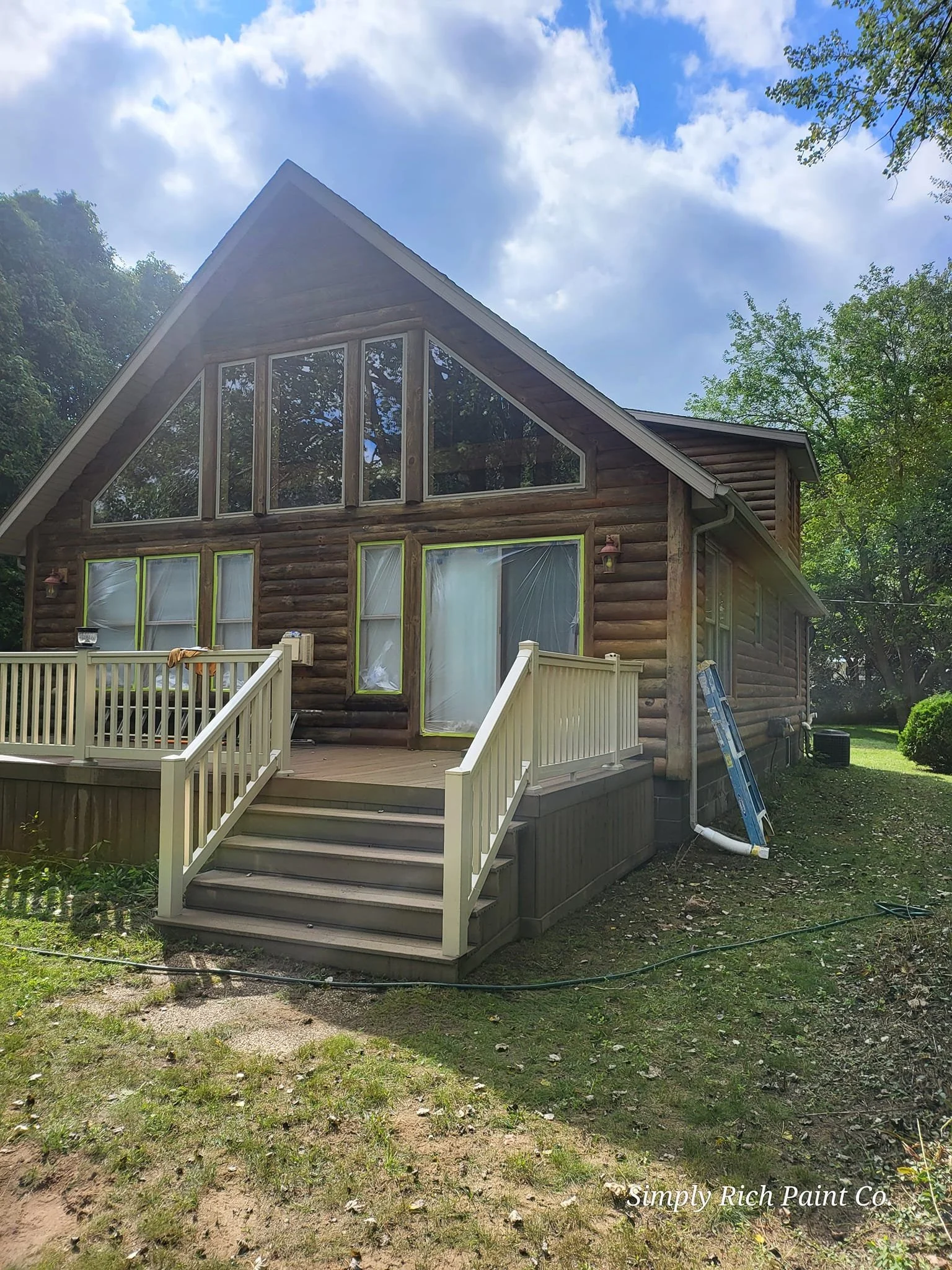 A wooden house with a steep A-frame roof, large glass windows, and a small porch with stairs, surrounded by trees on a sunny day.