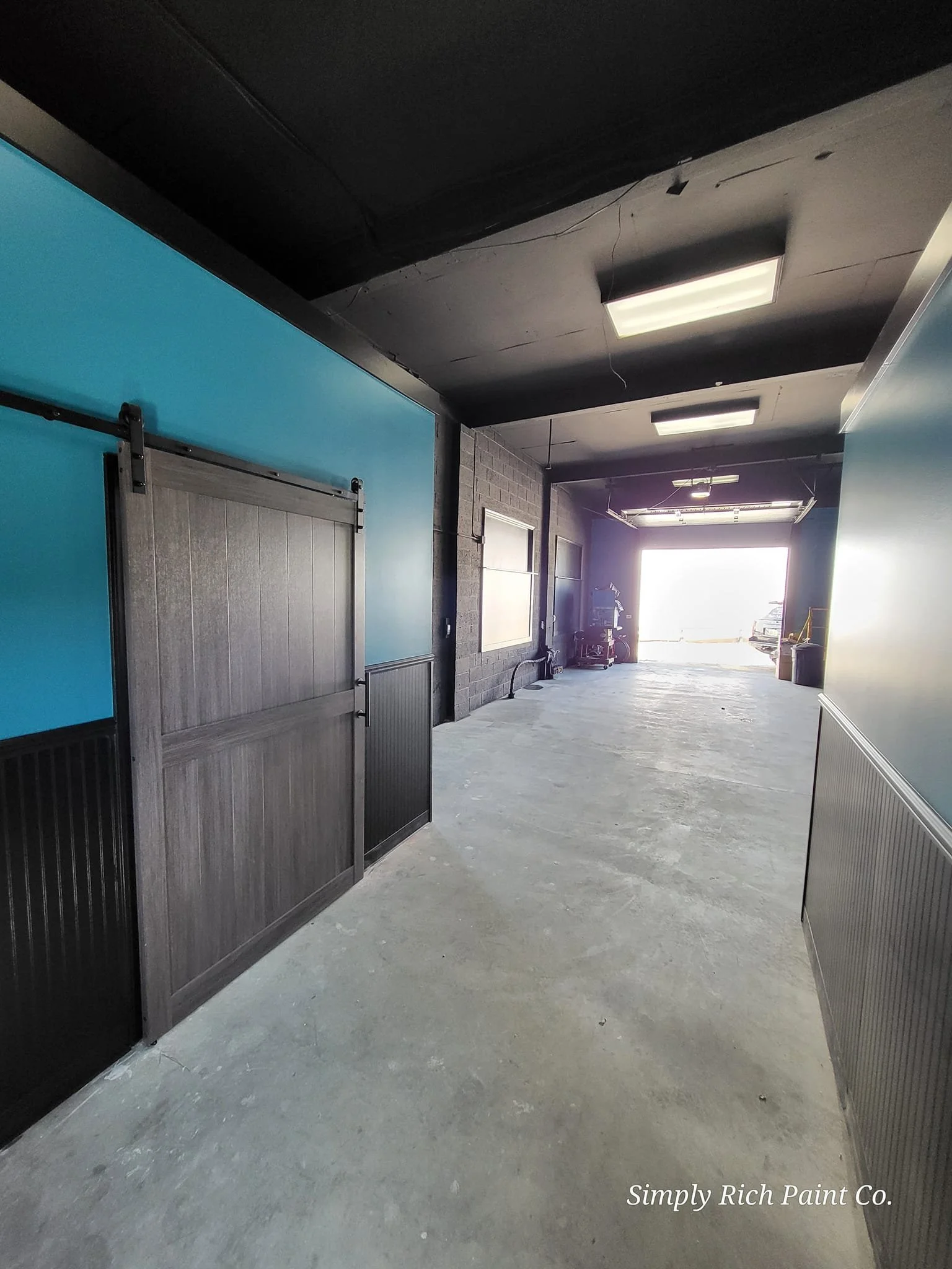 Empty garage with concrete floor, black ceiling, blue wall, and black-walled side. Garage door open, sunlight coming in, with a car partially visible outside. Interior has minimal items and some windows.