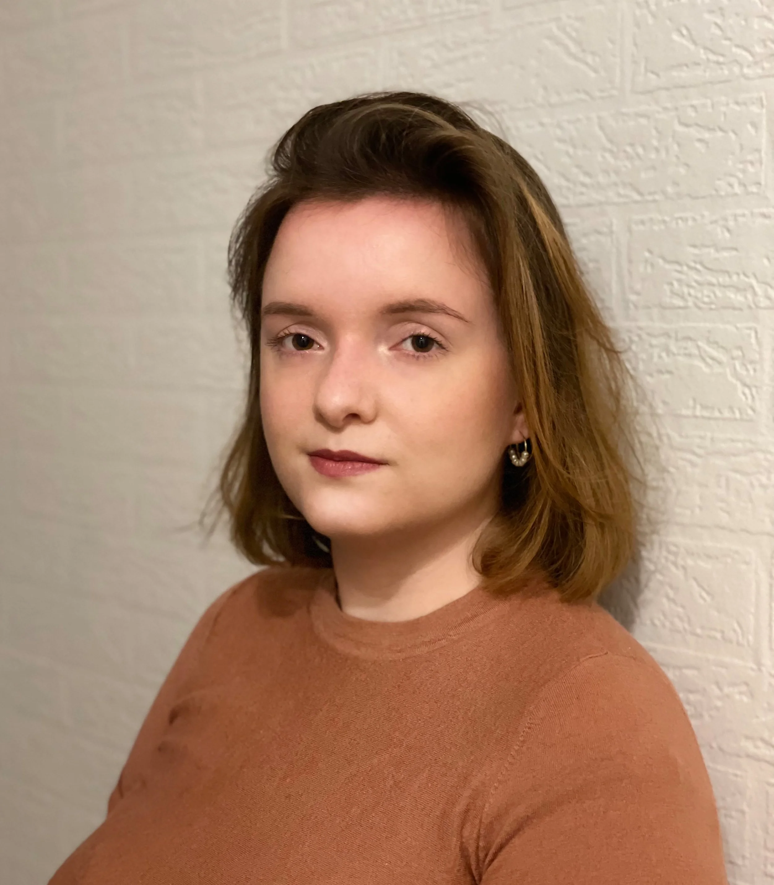 Porträt einer jungen Frau mit braunen Haaren, blassem Teint und dezenten Makeup, trägt ein braunes Oberteil, steht vor einer weißen Wand mit Struktur.