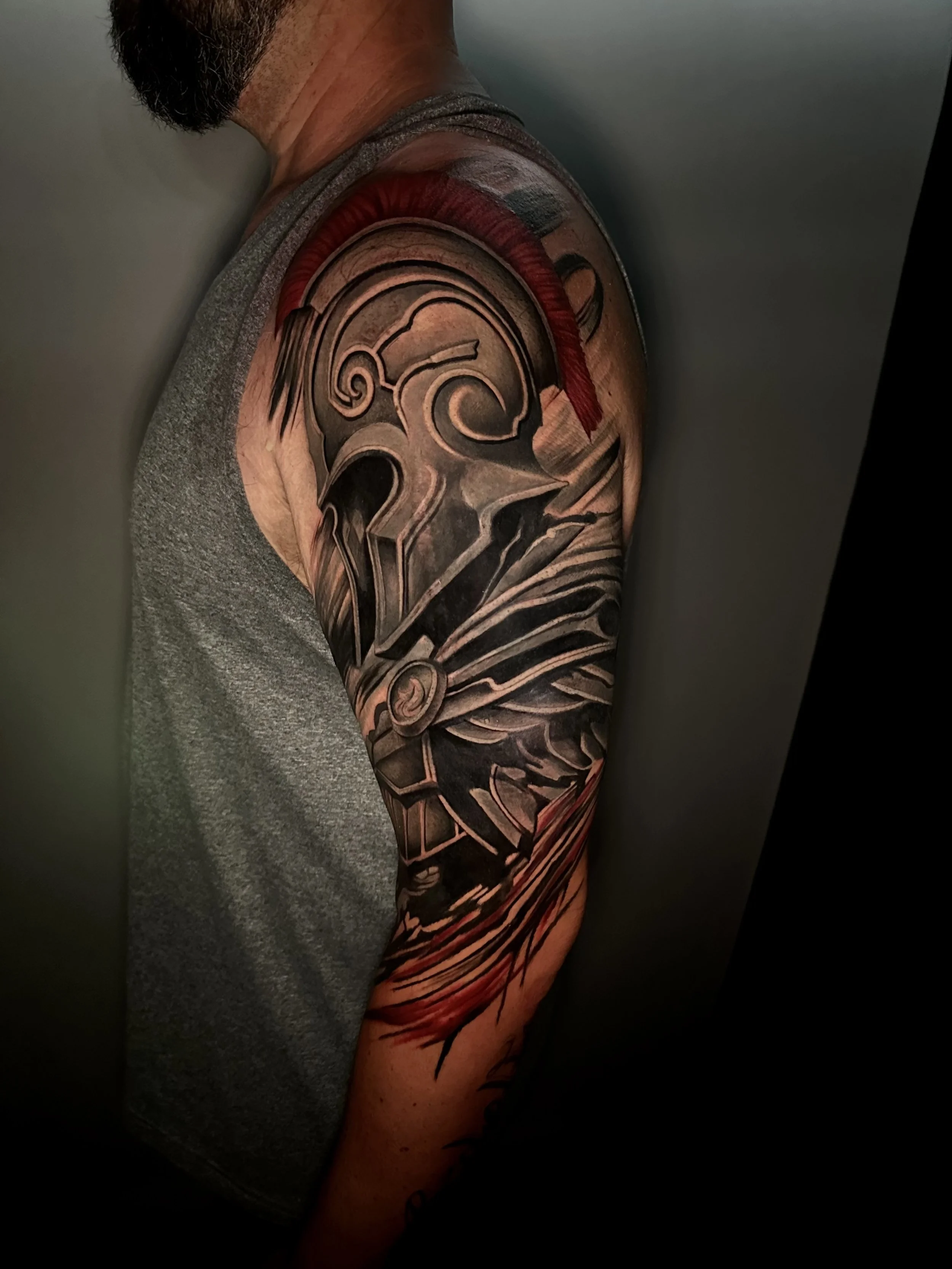 Coverup Black and gray tattoo of a Spartan helmet on a person's upper arm, with red and black accents and abstract design elements.