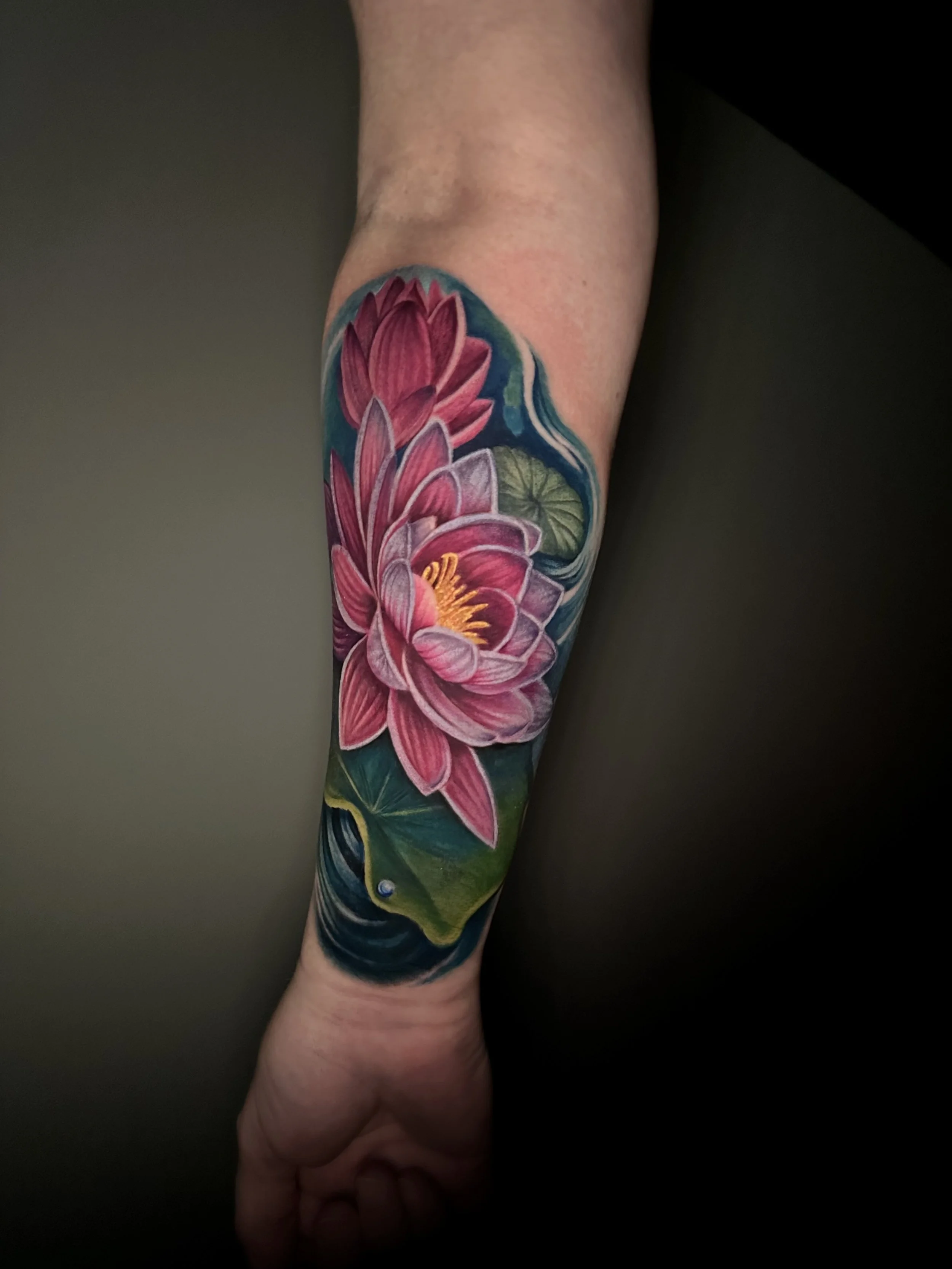 Colorful tattoo of pink water lilies and lily pads on a forearm against a dark background.