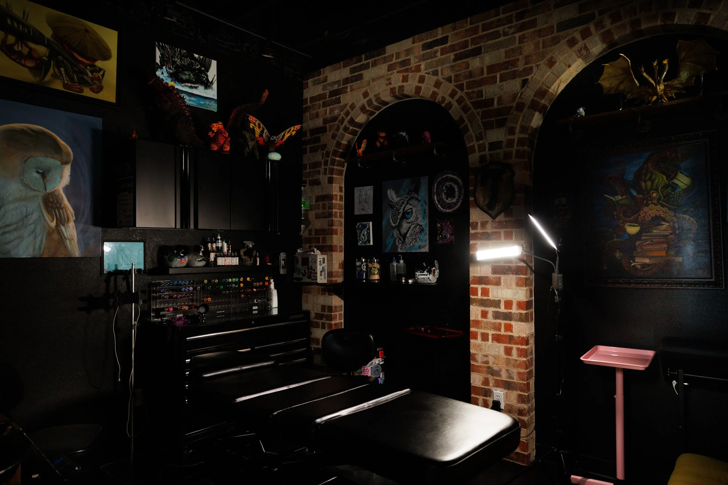 A tattoo studio with artwork, a tattoo chair, and various art supplies, with exposed brick walls and dark decor.