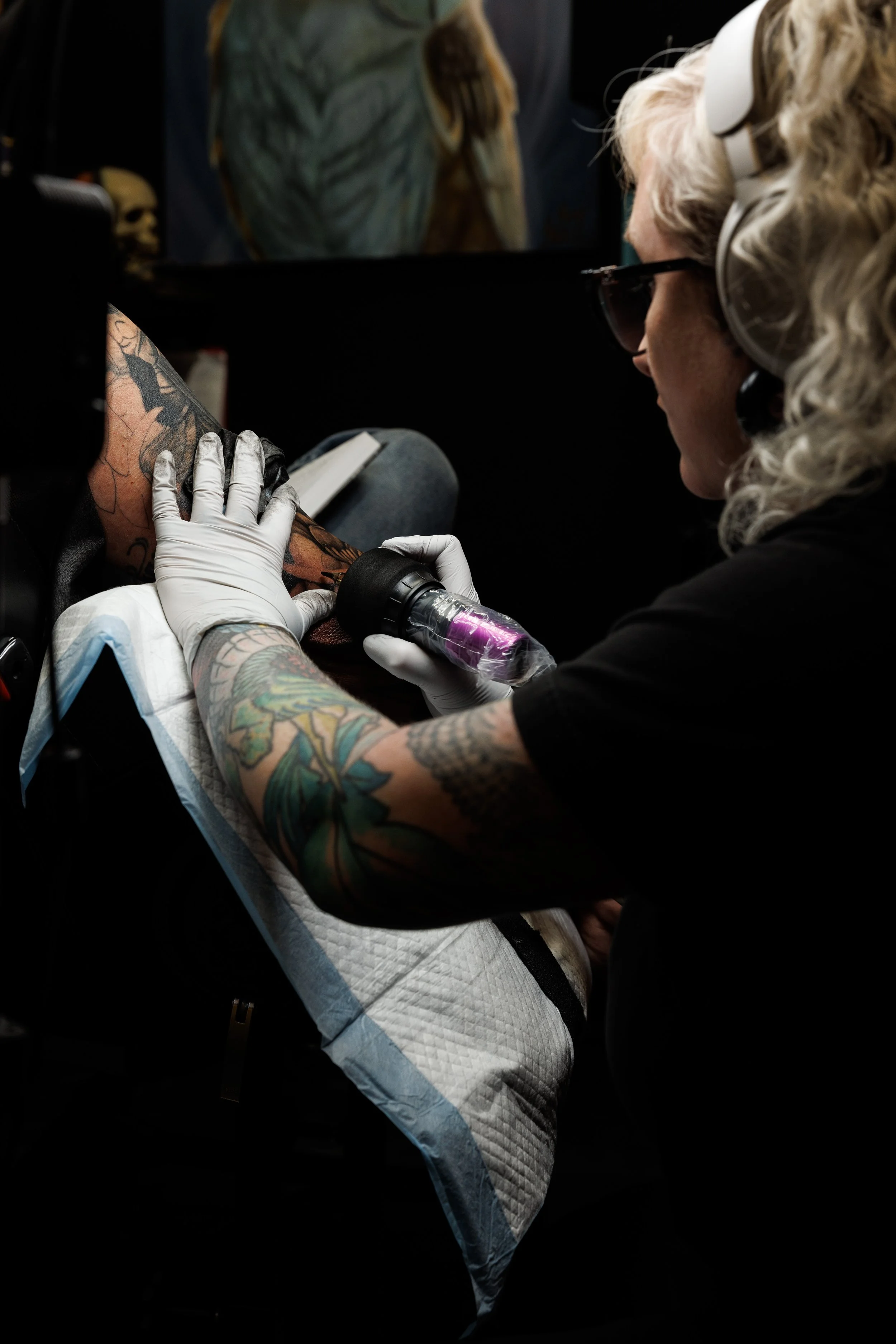 Tattoo artist in black shirt, with curly blonde hair, wearing glasses, white gloves, using tattoo machine on client's arm.