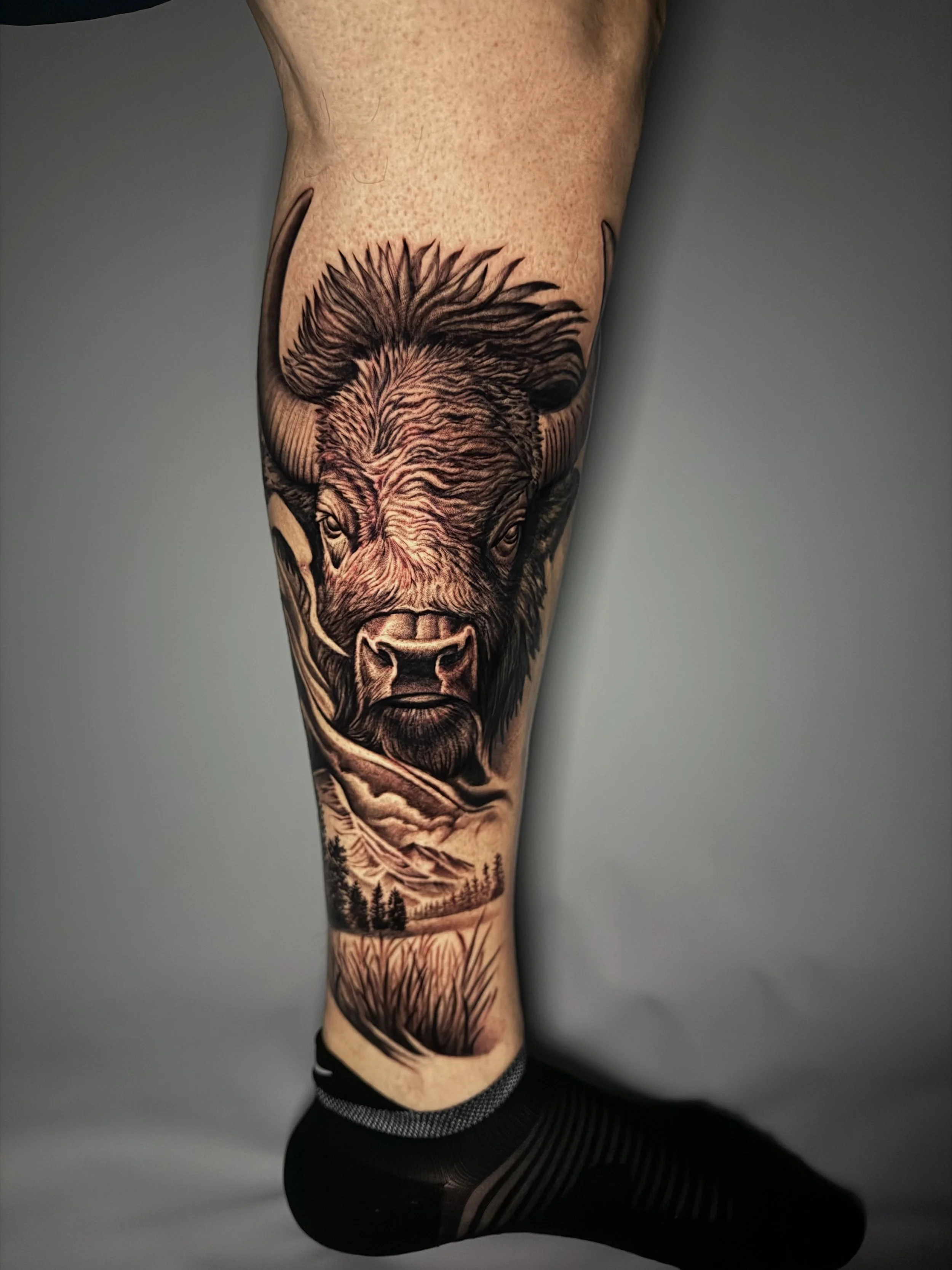 Tattoo of a realistic bison's face on a person's calf, set against a mountainous landscape background with trees and grass.