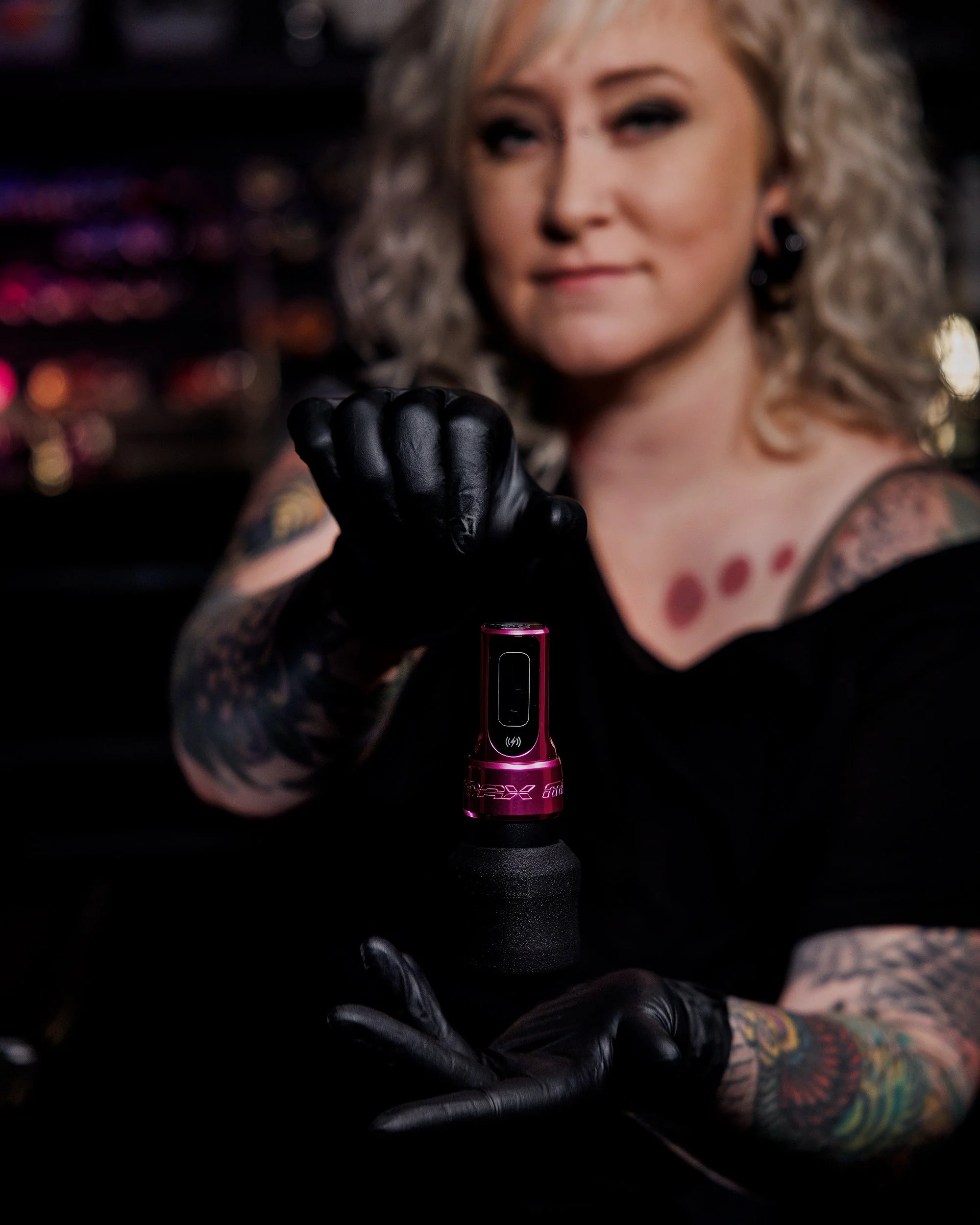 A woman with curly blonde hair and tattoos on her arms, wearing black gloves, is holding a pink vape or electronic cigarette in her hand.