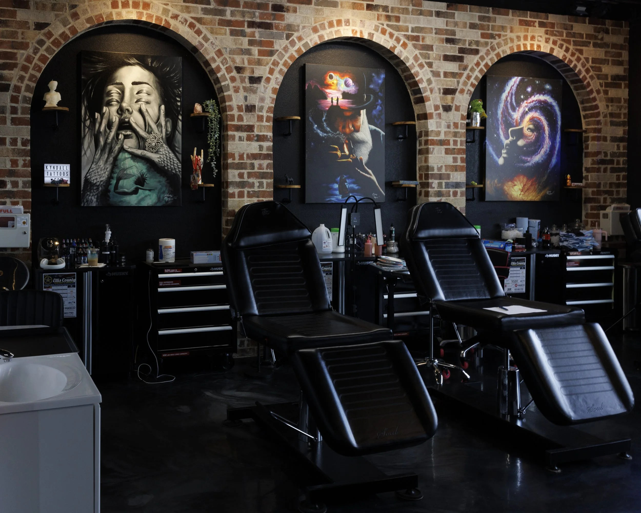 Tattoo studio with black tattoo chairs, art paintings on brick wall, and tattoo supplies on side tables.