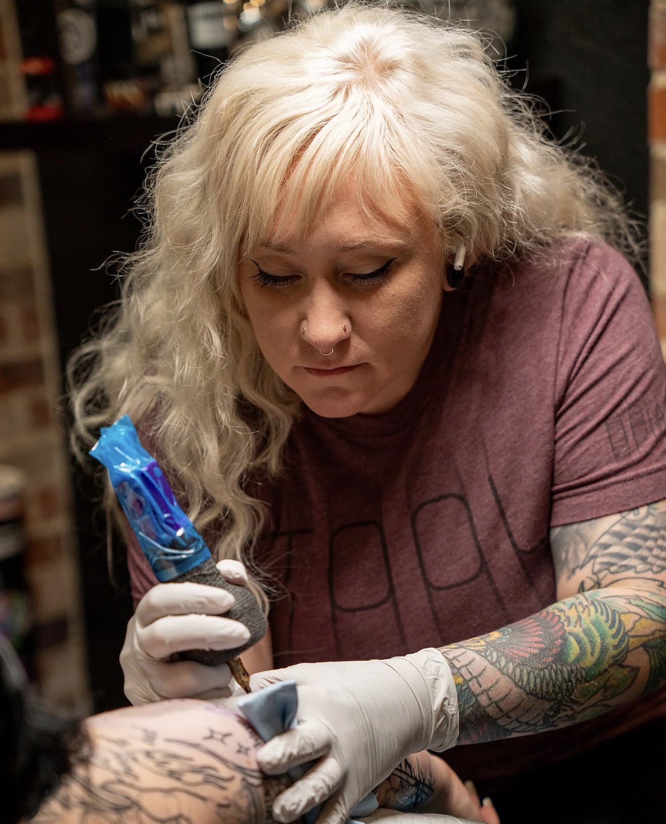 Tattoo artist with blonde curly hair and tattoos on her arms work on a client's arm, wearing gloves, in a tattoo studio.