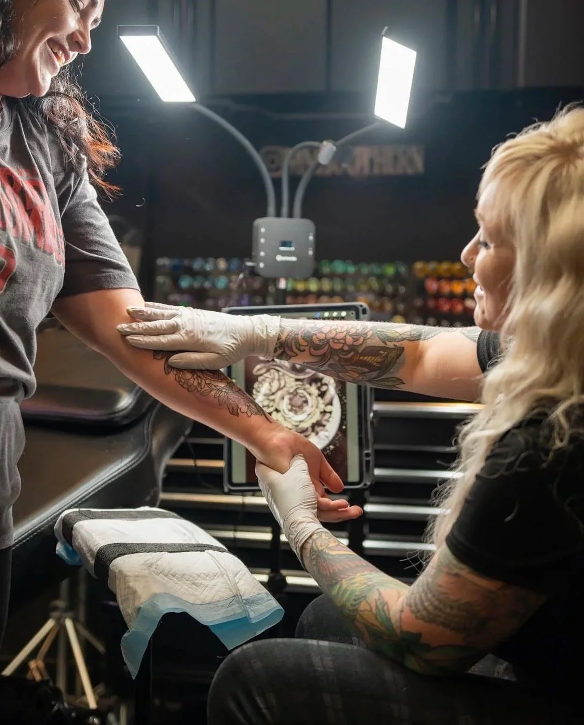 Tattoo artist working on a client's arm while smiling in a tattoo studio, with tattoo supplies and equipment visible in the background.