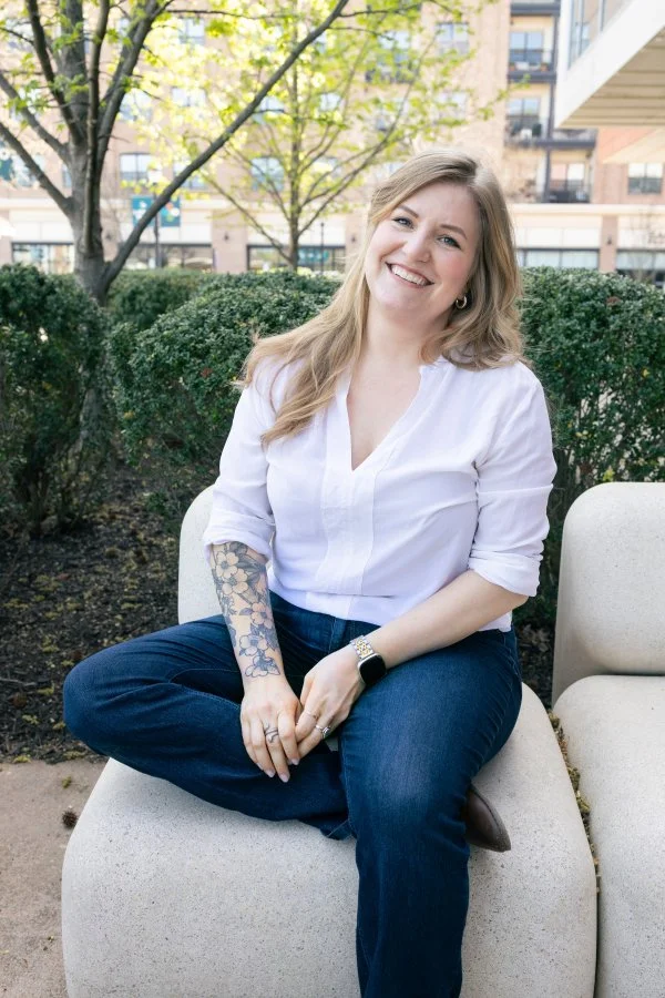 Jana Burtner, Designer & Self-Publishing Guide – A woman smiling, sitting cross-legged on a curved outdoor bench in a park, wearing a white blouse with sleeves rolled up to reveal dogwood tattoos, with greenery and buildings in the background.
