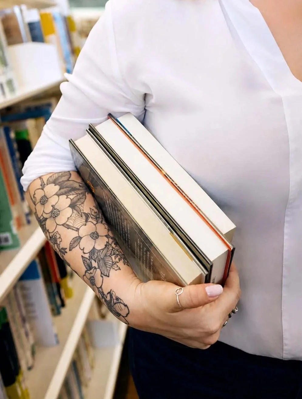 Person holding three books in a library, wearing a white shirt with rolled-up sleeves, displaying published works in the secure grasp of a supportive expert.