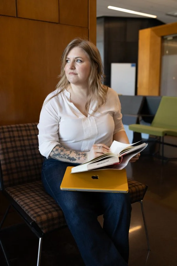 Jana Burtner, Designer & Self-Publishing Guide – A woman with light hair and tattoos sitting with a book to review, reading a notebook with a yellow laptop on her lap, in a modern interior space.