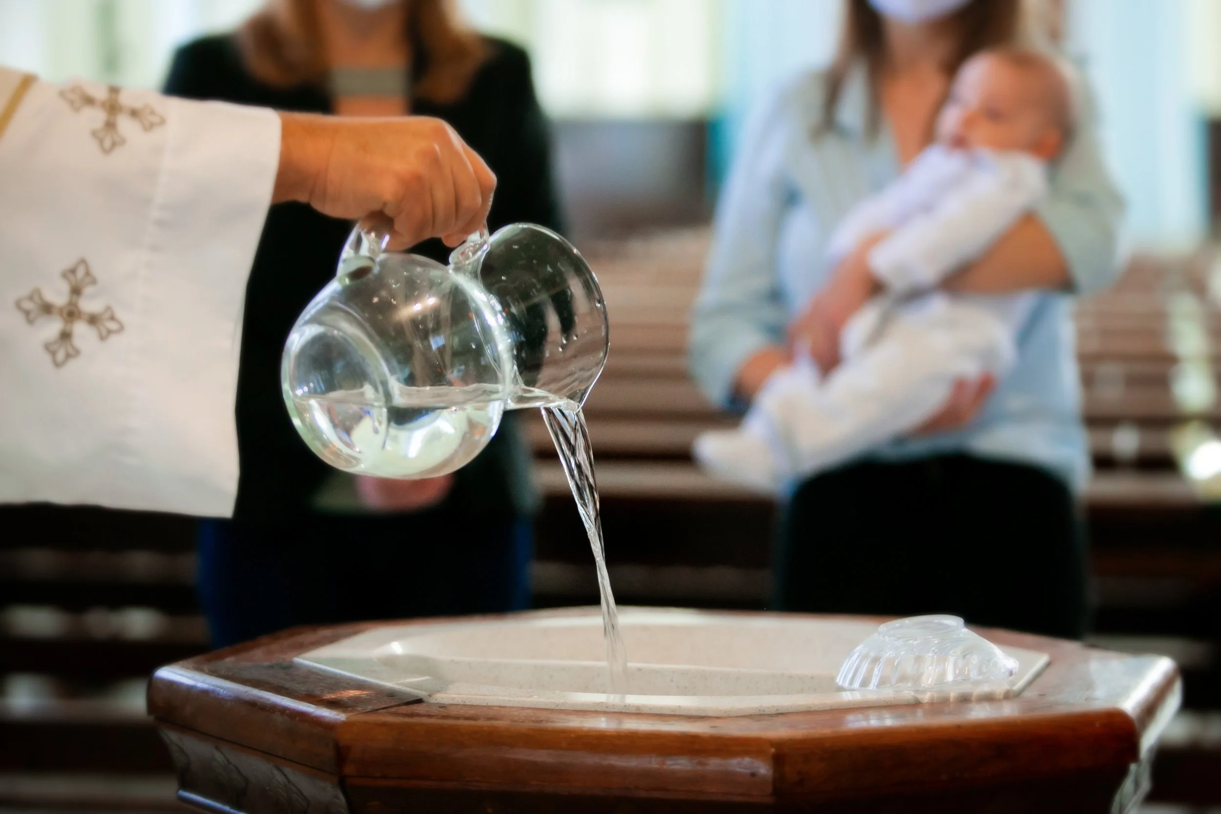 Pfarrer gießt Wasser ins Taufbecken. Im Hintergrund Täufling auf dem Arm der Mutter.