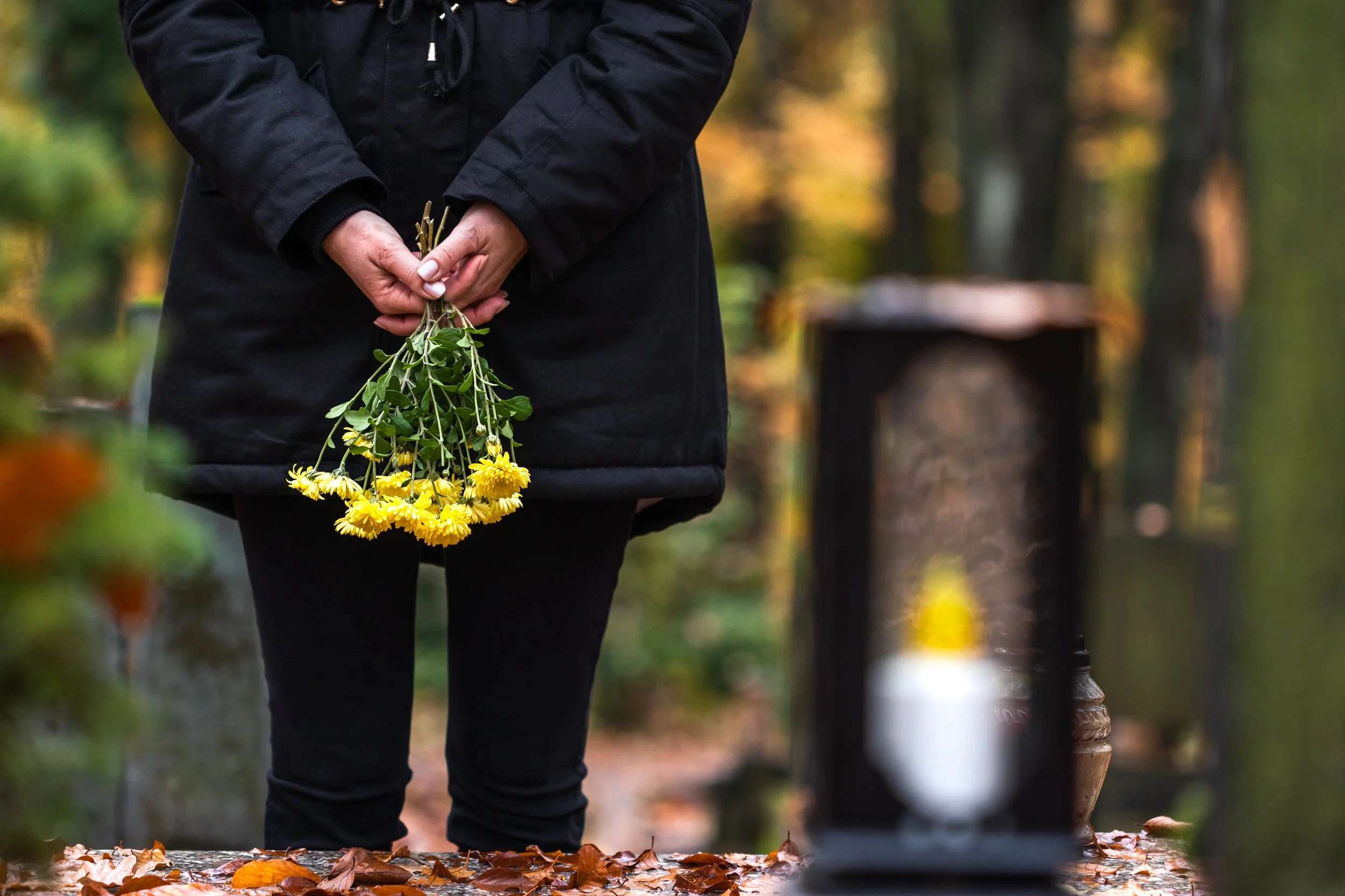Person in schwarzer Kleidung steht vor einem Grab mit Grabkerze und hält mit gefalteten Händen gelbe Blumen.
