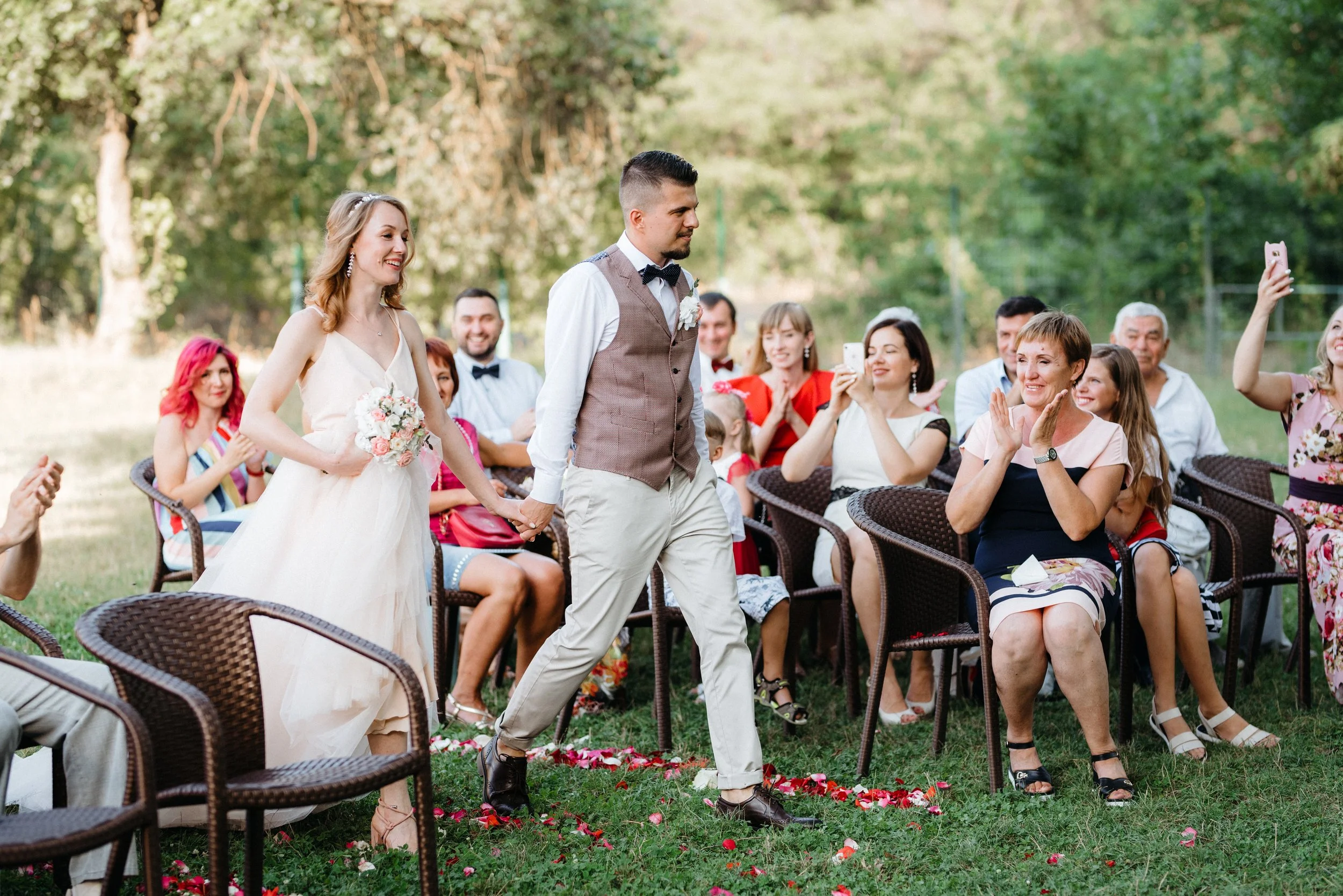 Hochzeitspaar geht Hand in Hand zur Trauung im Freien zwischen applaudierenden sitzenden Gästen hindurch.