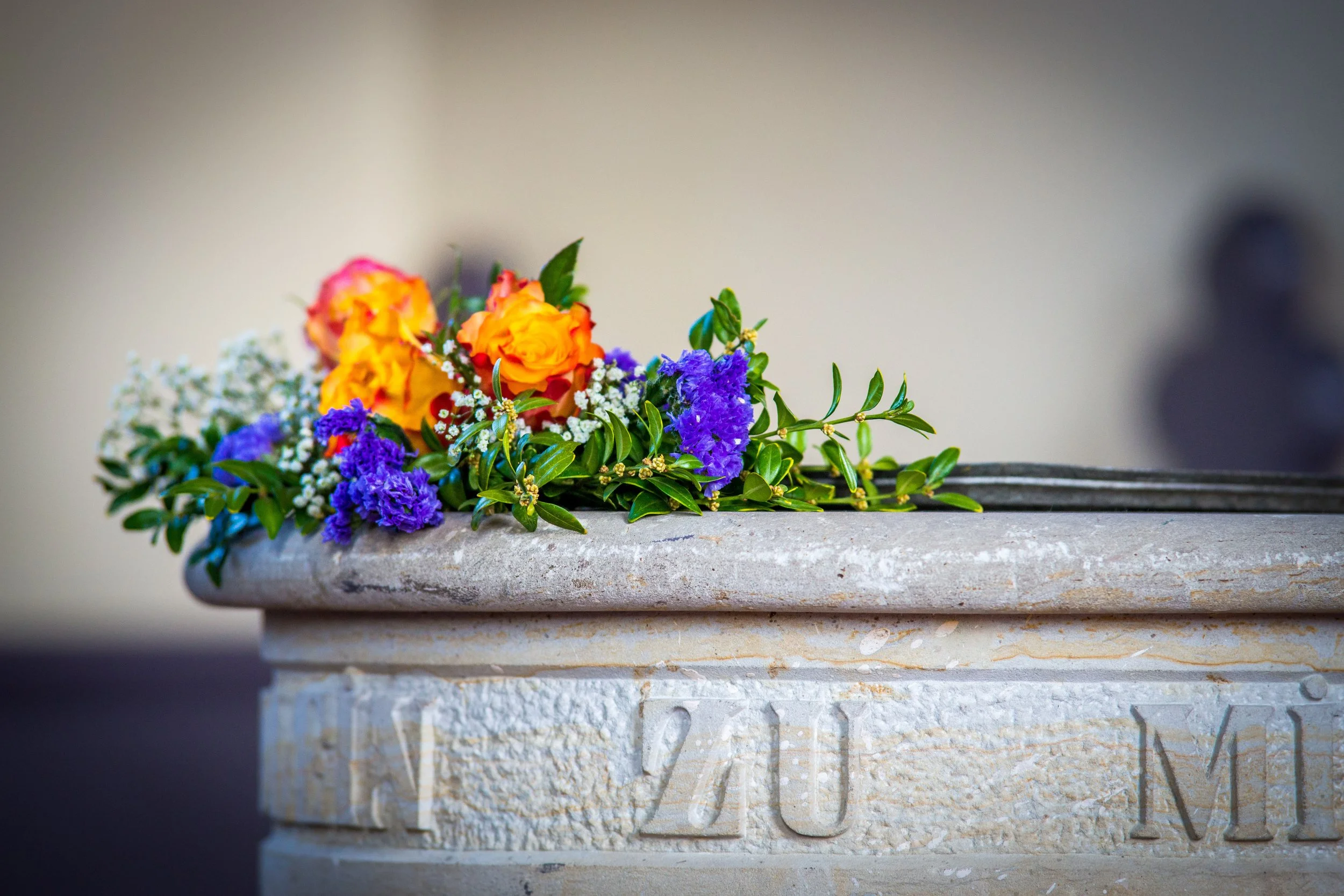 Detail eines Taufsteins, auf dem ein orange-violettes Blumengesteck liegt.