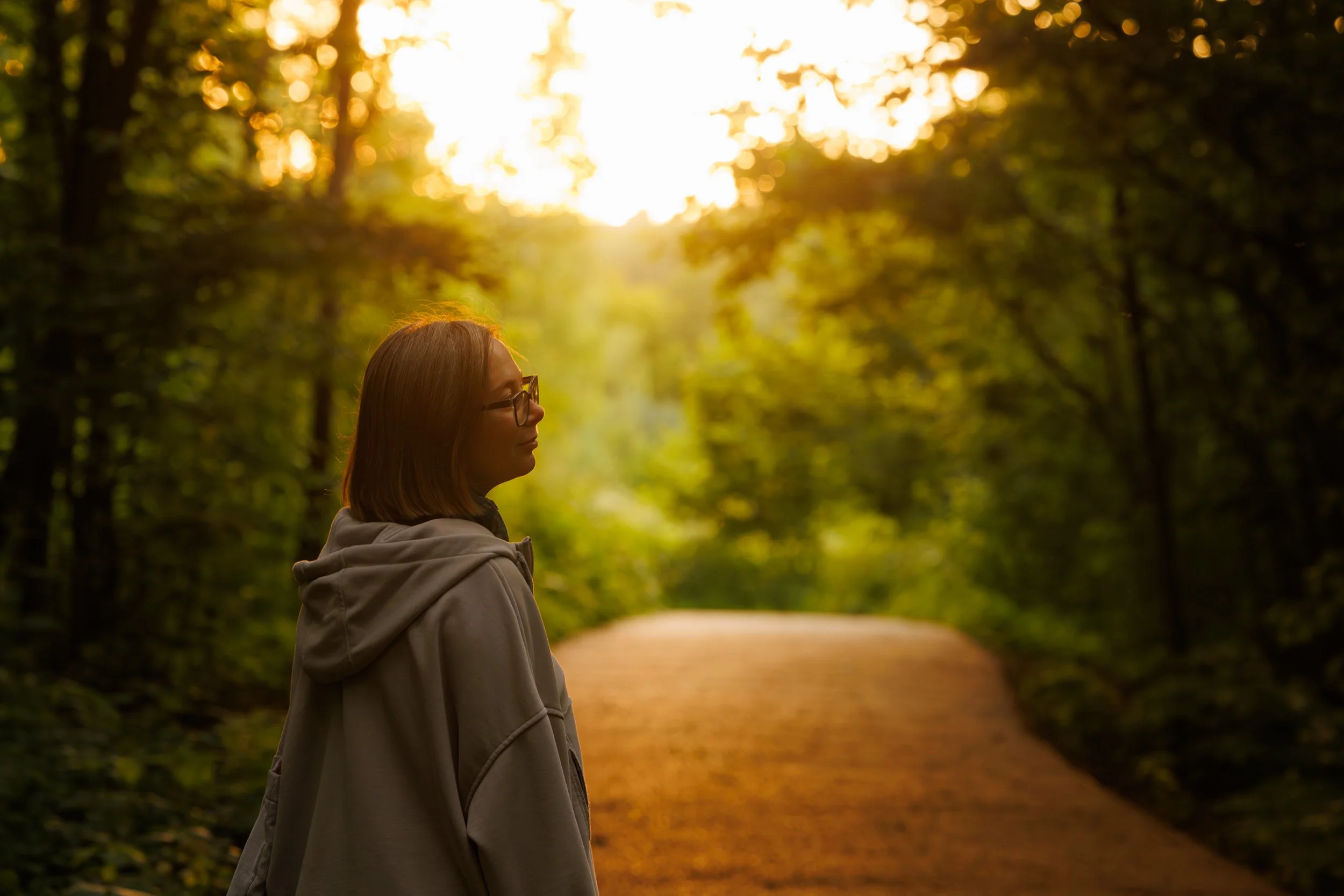 Eine etwa 50jährige Frau steht mit dem Rücken zur Kamera am Rande eines Waldwegs. Sie blickt gedankenverloren in die warm beleuchtete Landschaft.