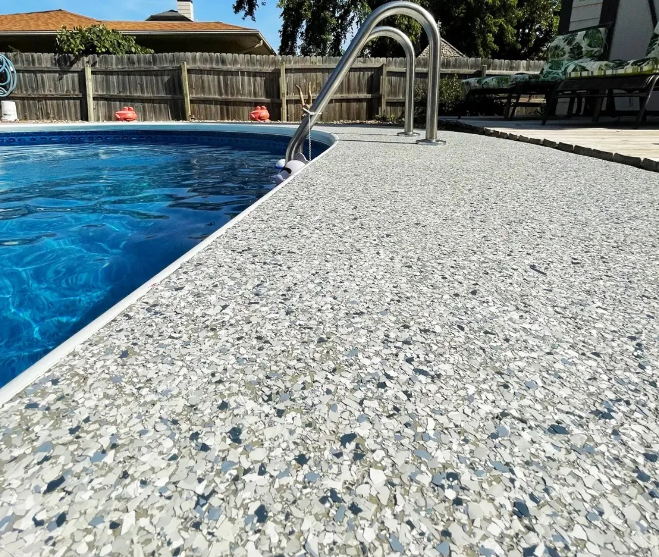 Close-up of a backyard swimming pool with a textured, speckled epoxy deck and metal handrail, surrounded by a wooden fence and outdoor furniture.