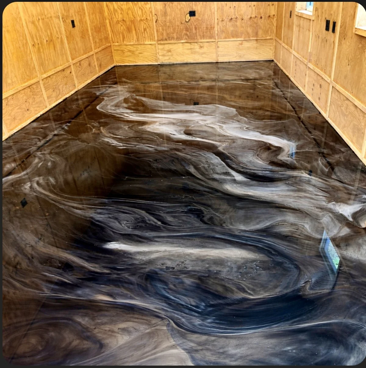 Room with plywood walls and a marbled epoxy floor, black surface with white and gray swirling patterns, resembling marble or fluid art.