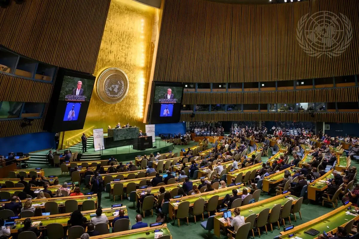United Nations General Assembly hall with delegates seated, large screens showing speakers, and the UN emblem on the wall
