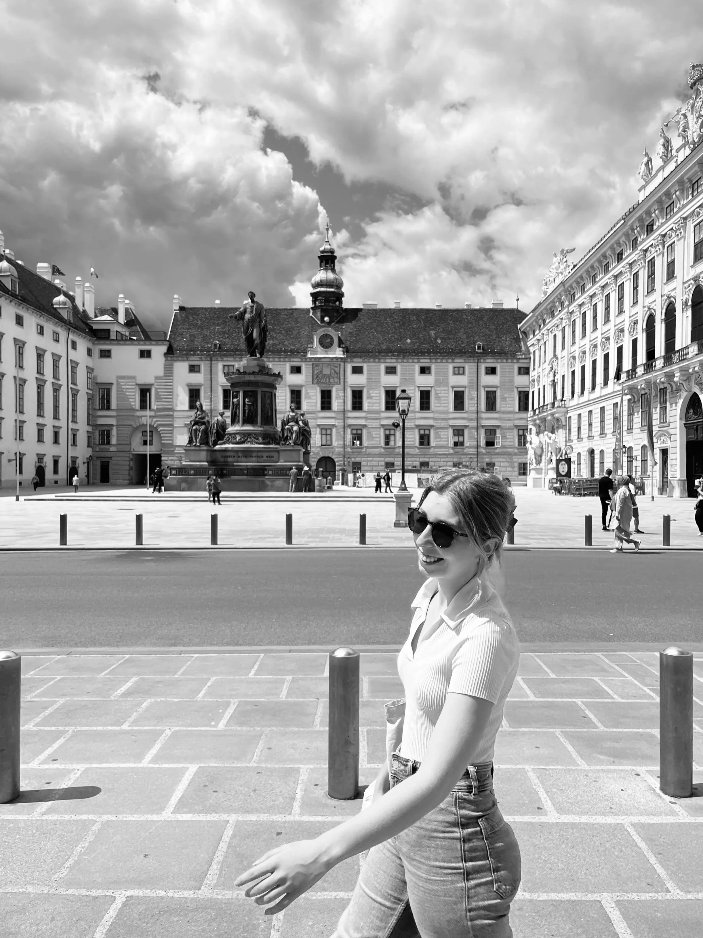A woman in sunglasses and a short-sleeve shirt walking on a city street with historic building and a statue in the background, sky with clouds.