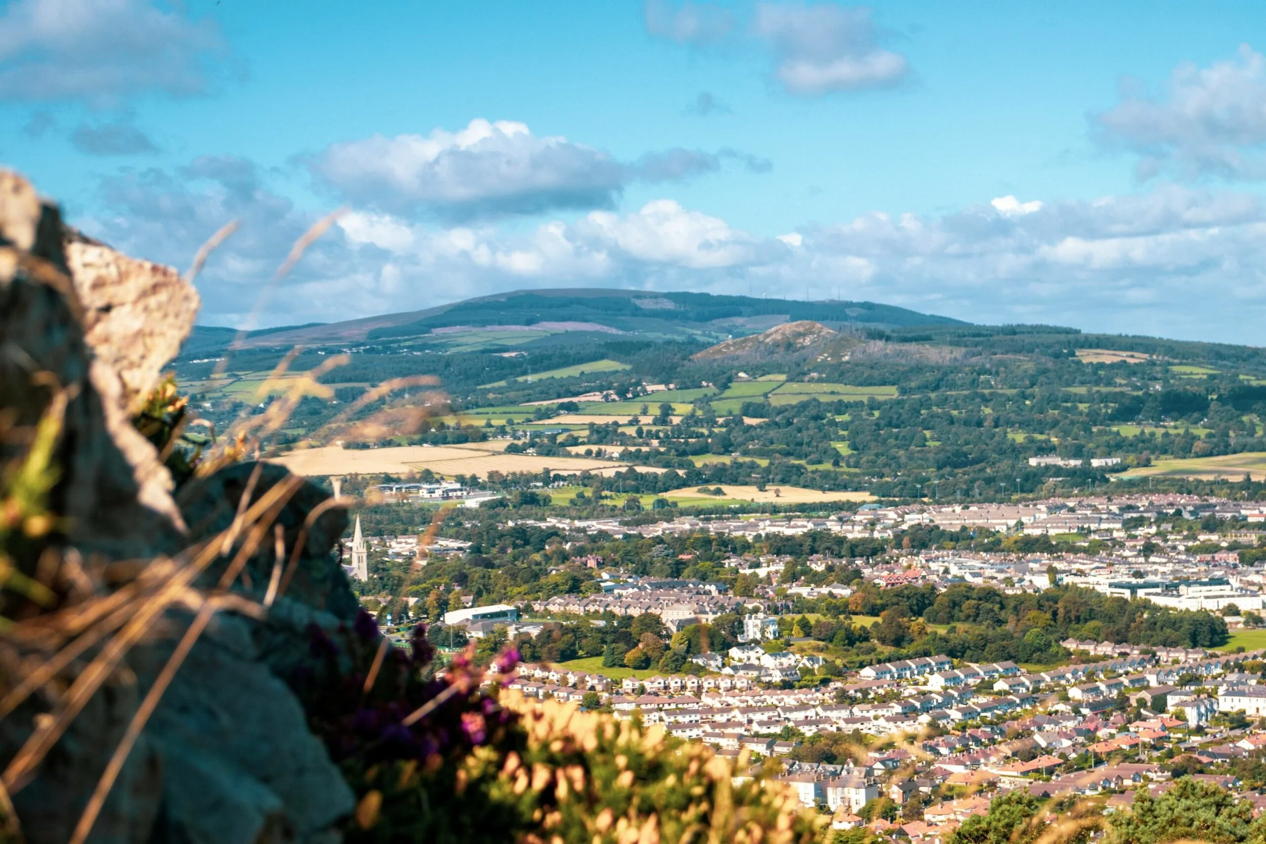 A panoramic view of an Irish town with a mixture of residential and commercial buildings, green fields, and rolling hills under a partly cloudy sky.