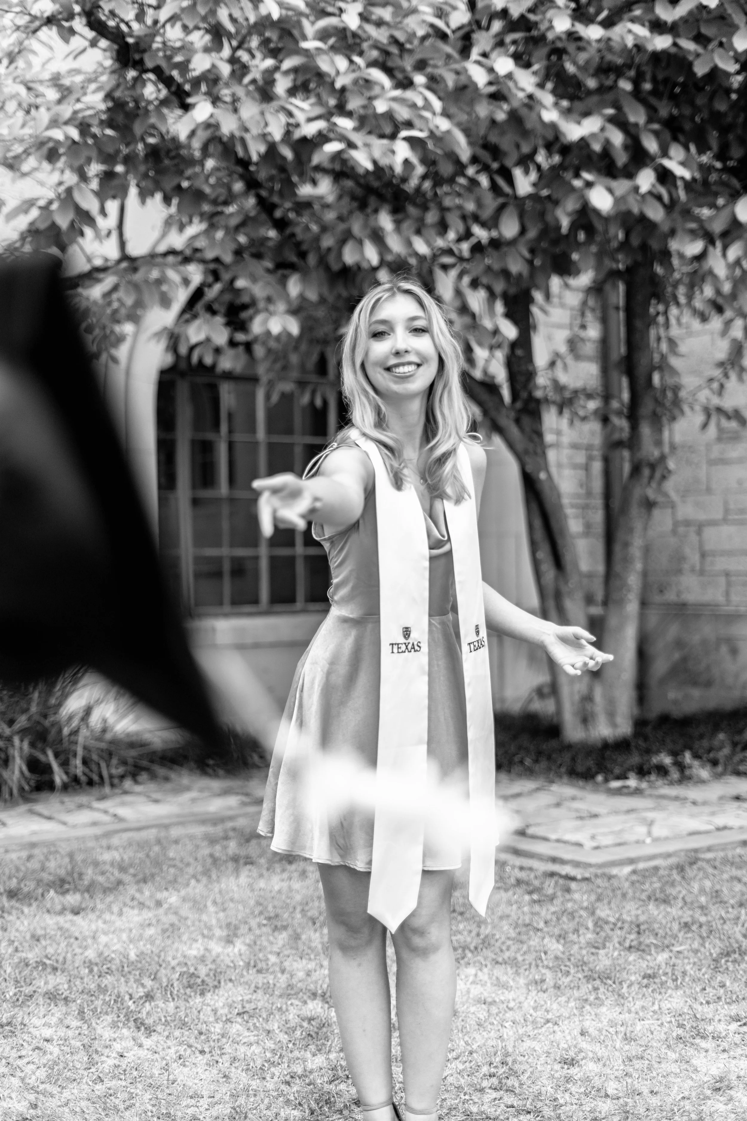A young woman in a graduation gown and stole standing outdoors with her arms open and smiling, with a tree and building in the background.