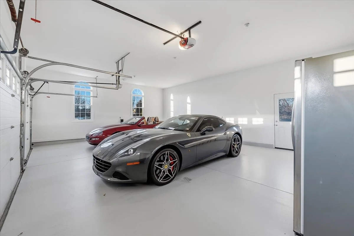 A garage interior with two luxury sports cars, a gray Ferrari and a red convertible, parked inside. The garage has white walls, a white floor, and large windows letting in natural light.