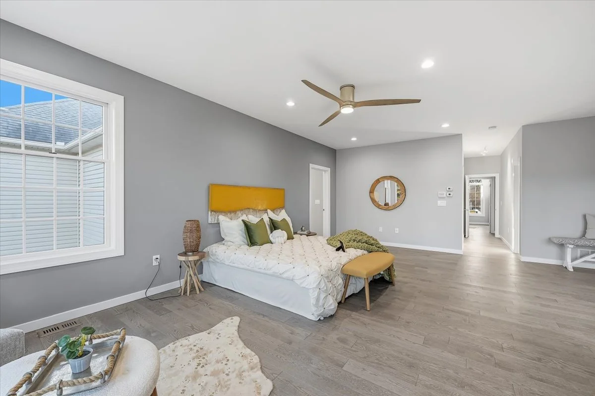 A spacious bedroom with gray walls, a large window, a bed with white and green pillows, a yellow headboard, a bench at the foot of the bed, a small side table with a lamp, a round mirror on the wall, wood flooring, and a ceiling fan.