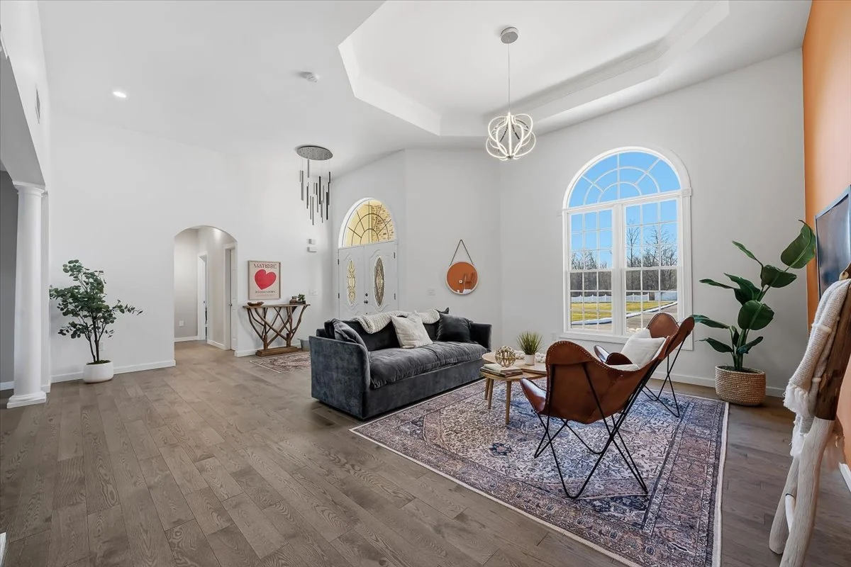 Bright living room with large arched window, white walls, wooden floors, modern light fixtures, black sofa, leather chairs, and indoor plants.