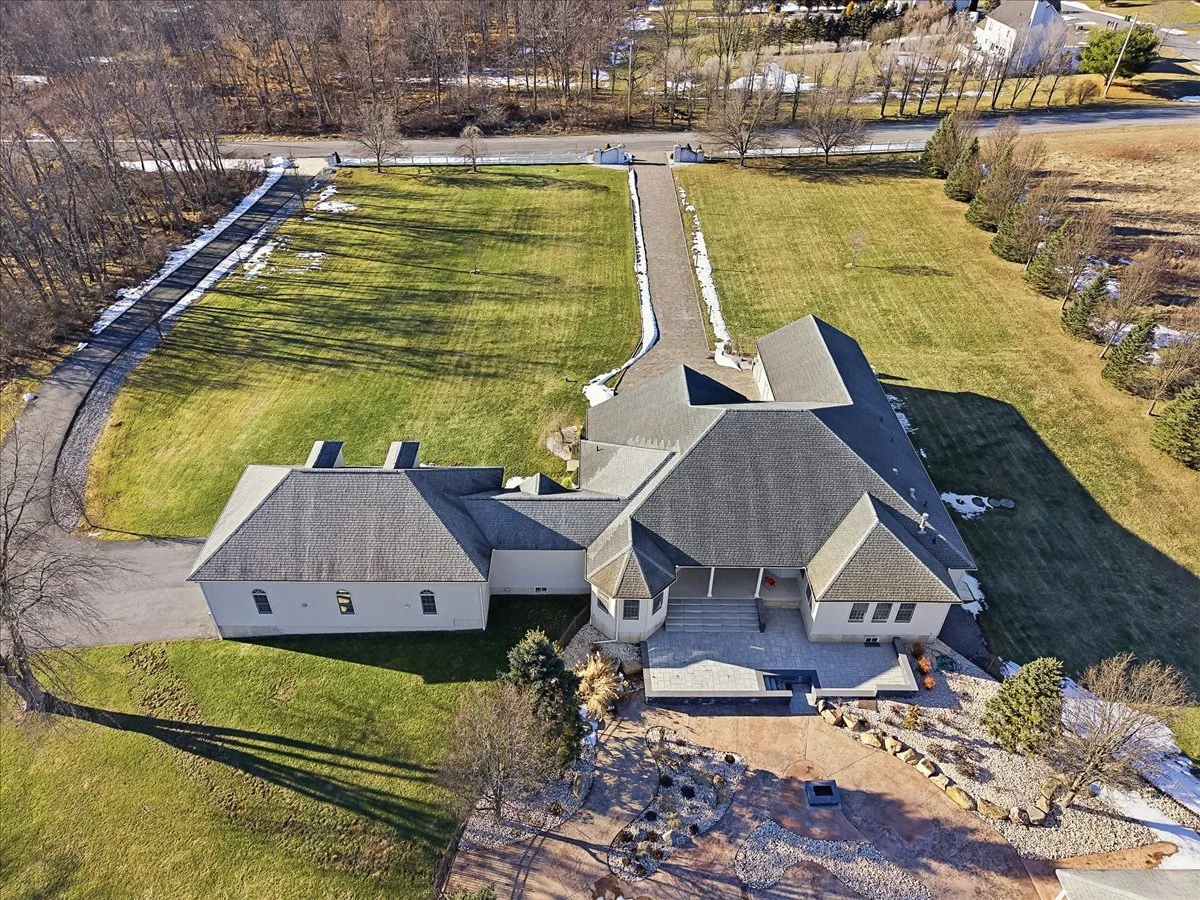 Aerial view of a large house with a gray roof, front porch, and a paved patio. The property has expansive green lawns, some patches of snow, and a gravel driveway. There are trees and bushes around the house, with a road and additional trees in the b