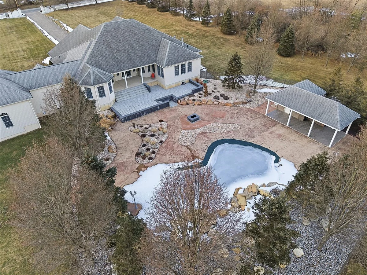 Aerial view of a backyard with a house, a small partially frozen pond, a covered patio, a fire pit, landscaped garden beds, and trees.