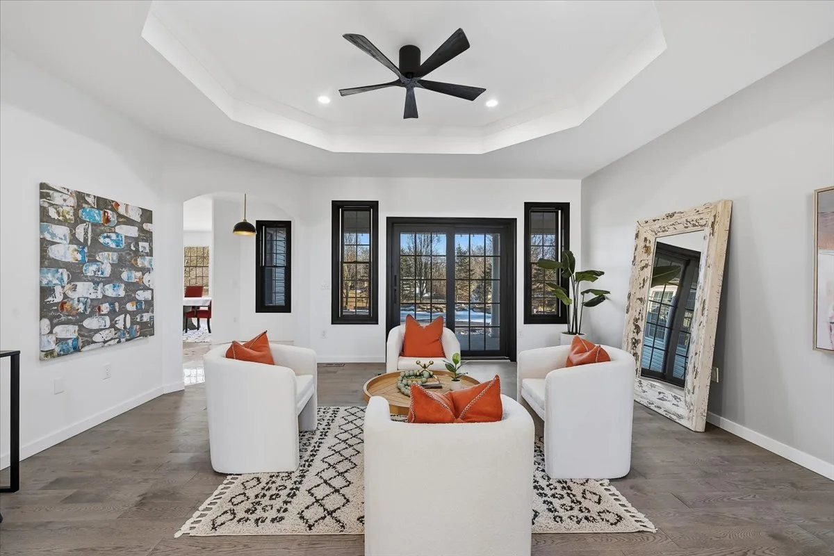 Living room with white chairs, an orange pillow on each, a round wooden coffee table, a patterned rug, black framed windows and sliding door, a large ornate mirror leaning against the wall, a potted plant, and modern artwork, with a ceiling fan overh