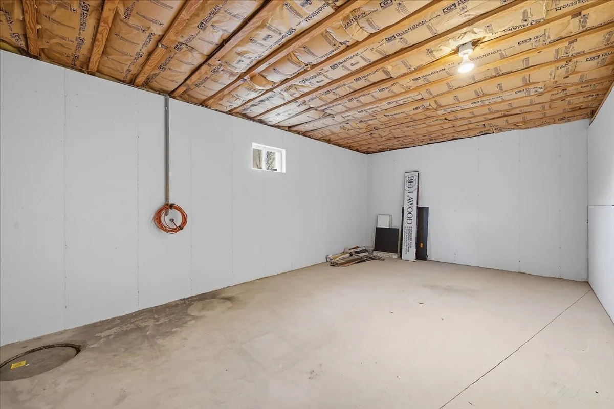 Empty room under construction with drywall walls, exposed ceiling insulation, and a small window. Tools and materials are stacked in one corner.