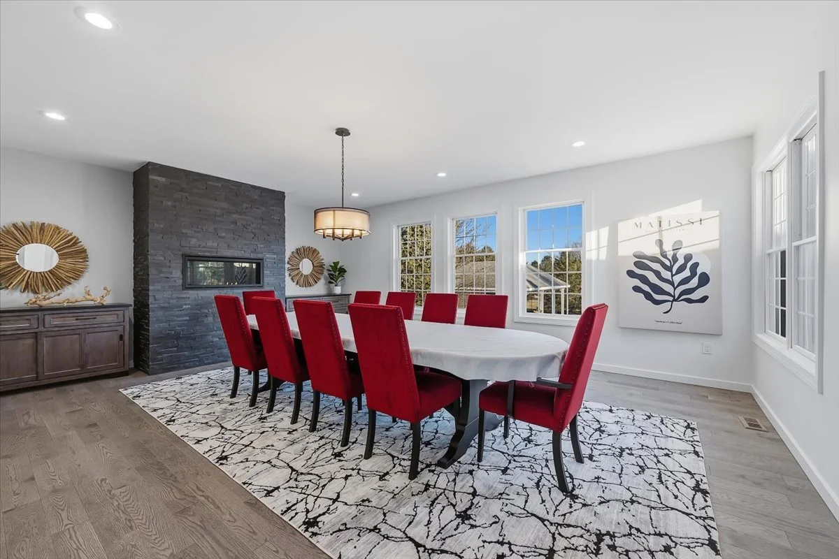 A dining room with a large table covered with a white tablecloth and ten red upholstered chairs. There are three windows, a black brick fireplace, a wooden sideboard, and decorative round mirrors on the wall. A large abstract art piece hangs on the r