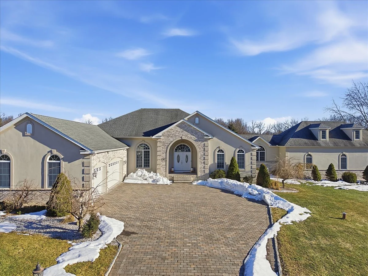 Front view of a large house with a brick driveway, snow on the ground, and a clear blue sky.