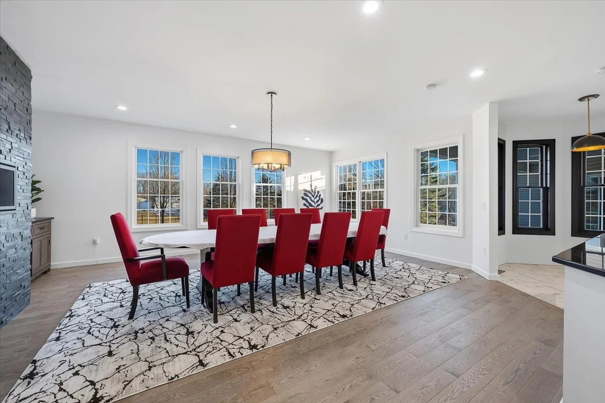 A modern dining room with ten red upholstered chairs around a large white rectangular table, a black and beige circular chandelier overhead, white walls, large windows showing a view of outdoors, and a patterned area rug on wooden floor.