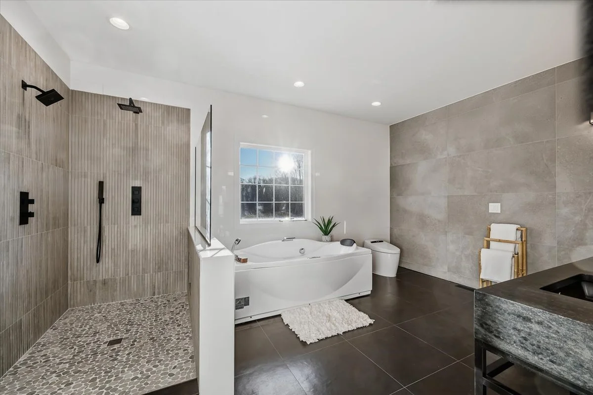 Modern bathroom featuring a walk-in shower with pebble floor, a bathtub, a window letting in natural light, a potted plant, a towel ladder, and neutral-colored tiles.