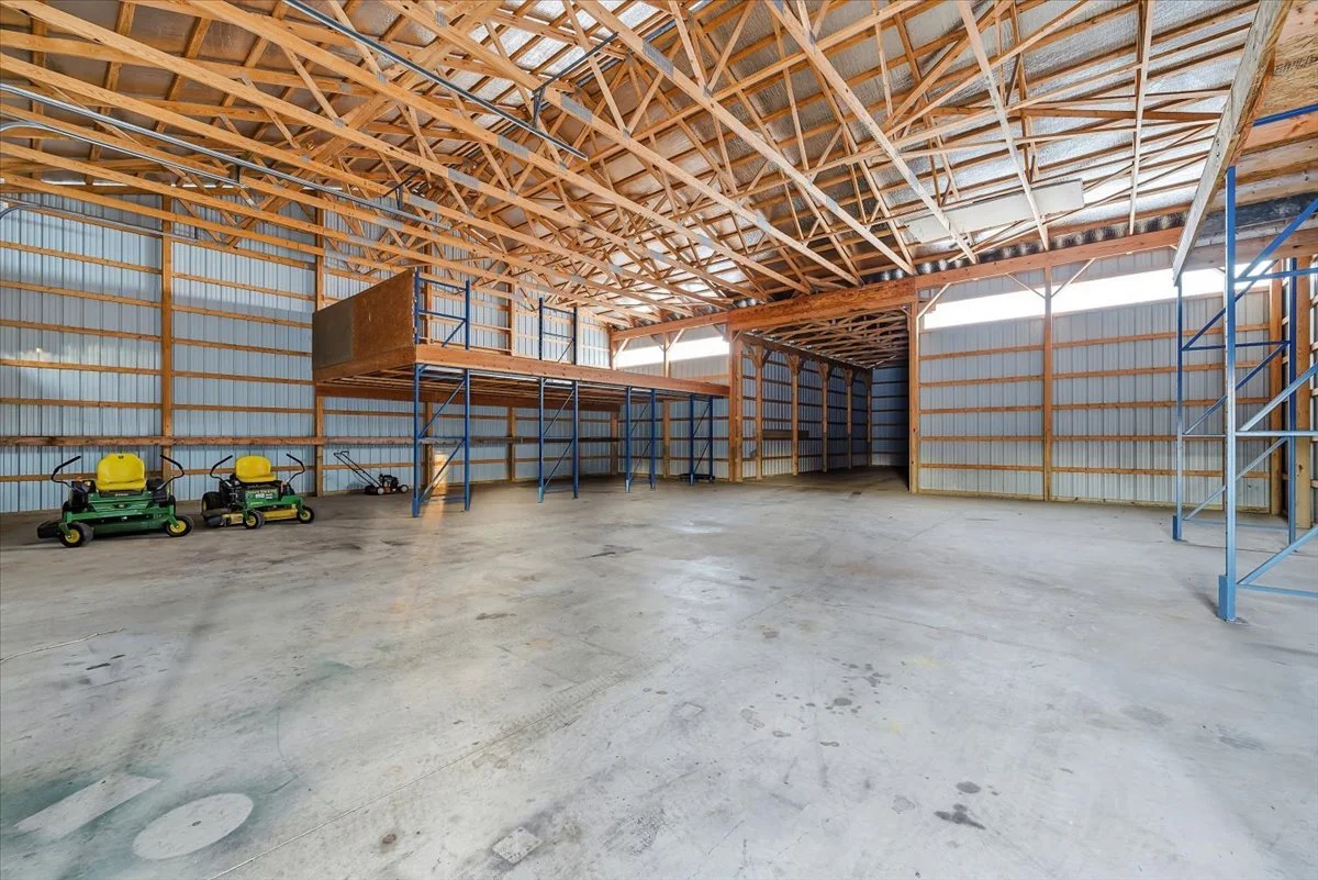 Empty metal building with unfinished wooden roof trusses, a mezzanine with metal framing, concrete floor, and two small green and yellow lawn mowers.