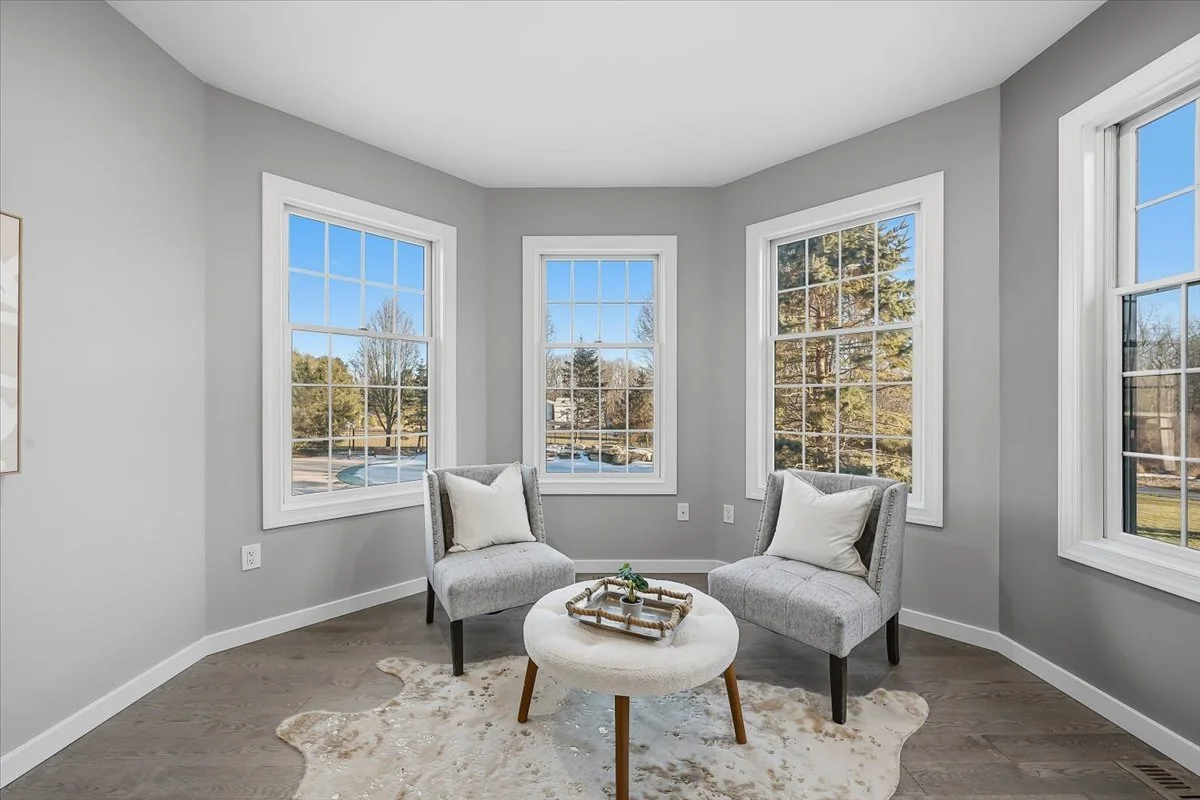 Sunlit corner of a modern living room with gray walls, three large windows, two gray armchairs with white pillows, and a small round ottoman with a tray and plant, placed on a light-colored cowhide rug.