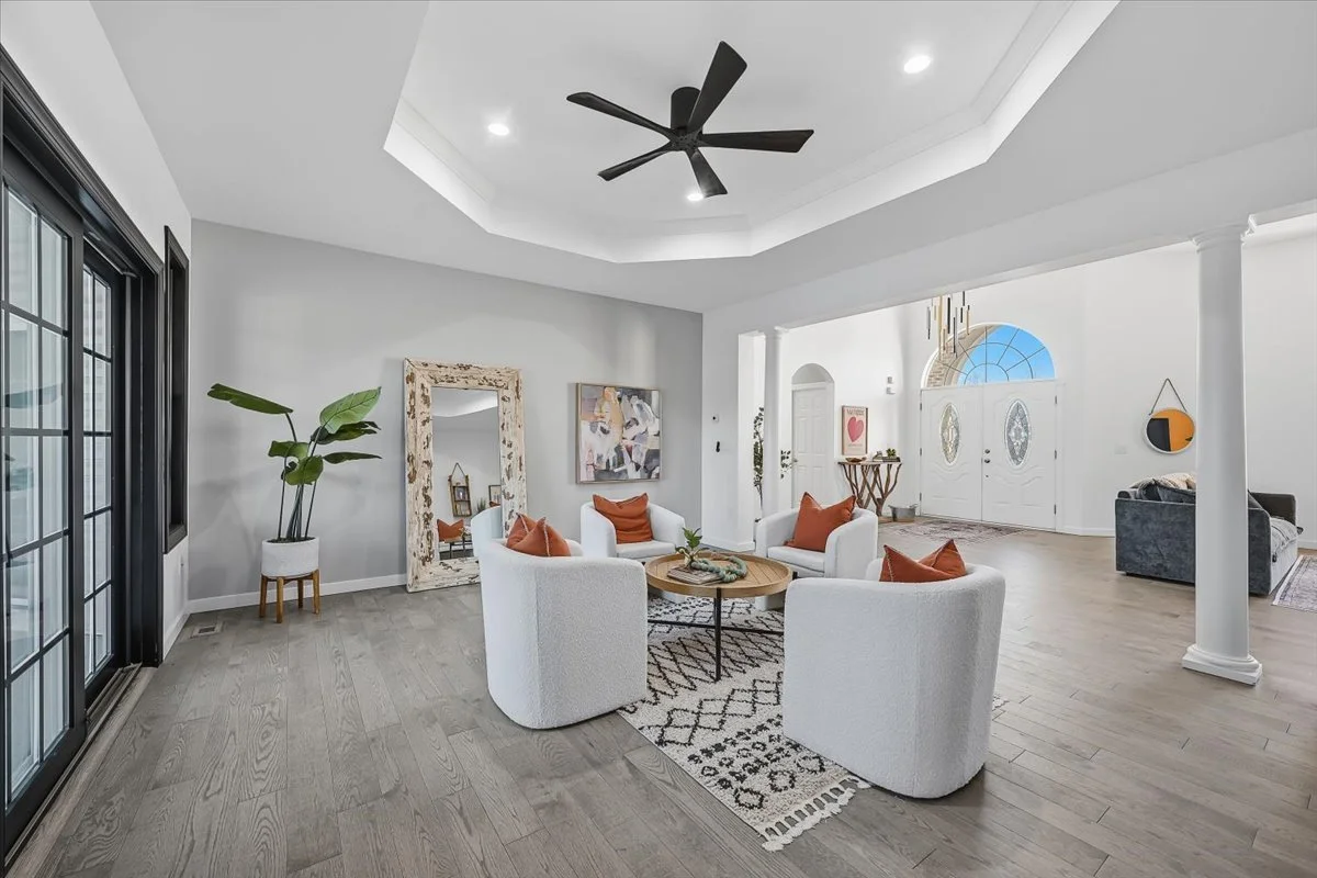 Living room with four white armchairs with brown pillows arranged around a round coffee table on a patterned rug, large mirror leaning against the wall, potted plant, and artwork on the wall. In the background, front door with a semi-circular window 