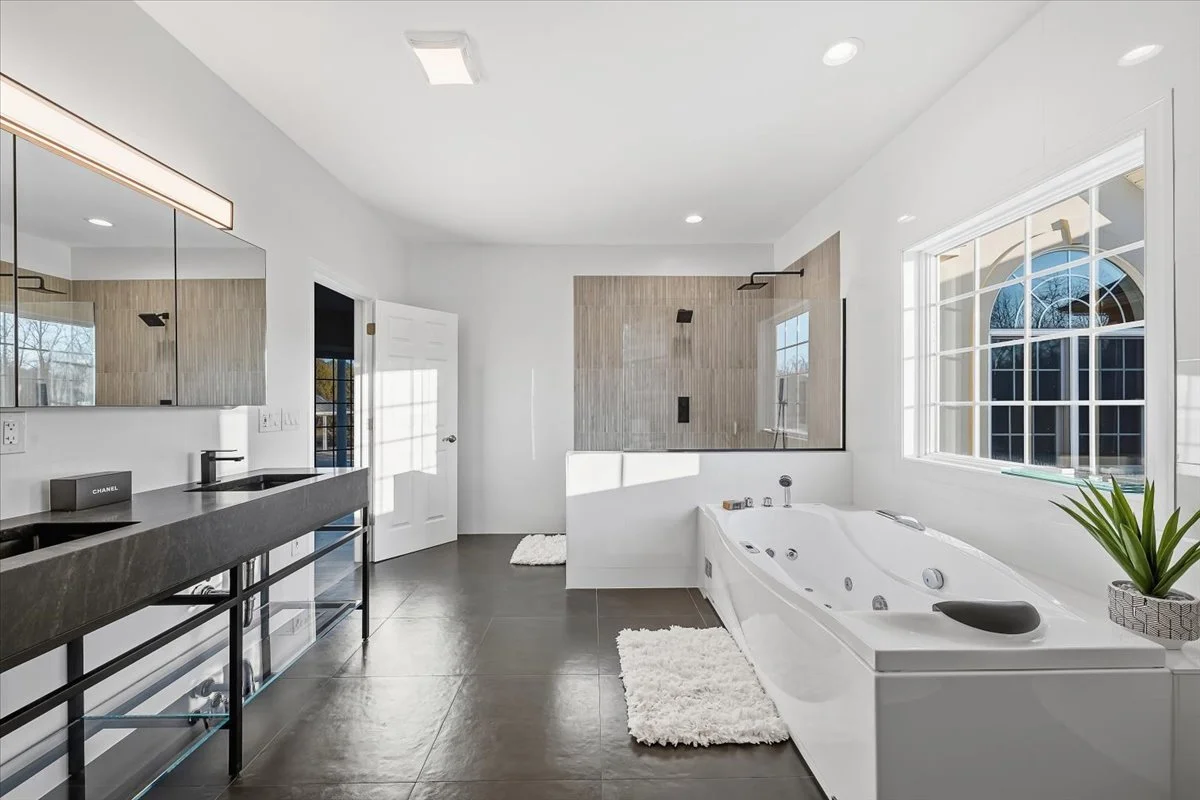 Modern bathroom with a large white whirlpool bathtub next to a window, wooden tiled shower area with a black showerhead, black double sink vanity with a mirror, and dark tile flooring.