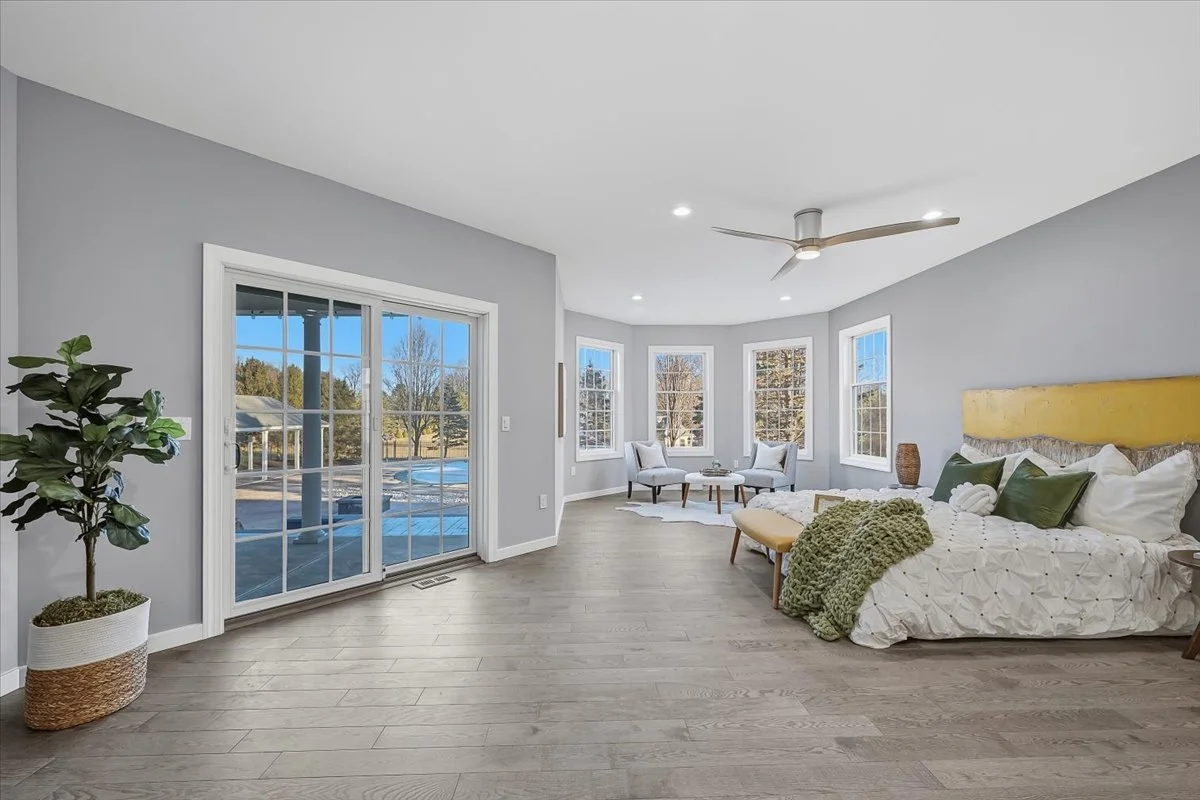 Bright bedroom with a large bed with green and white pillows, a green throw blanket, and a yellow headboard. There are two white chairs and a small table by the bay window, with a ceiling fan and hardwood floors.