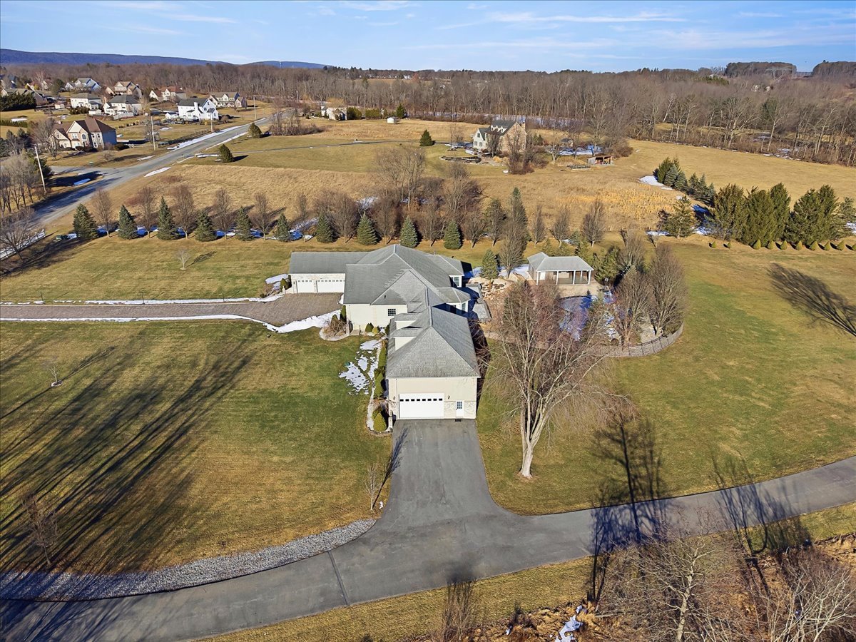 Aerial view of a large residential property with a house, driveway, garage, trees, and open fields in a suburban neighborhood.