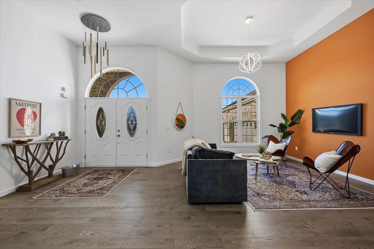 Living room with white and orange walls, hardwood floors, a black sofa, a chair, a TV mounted on the orange wall, and decorative lighting fixtures.