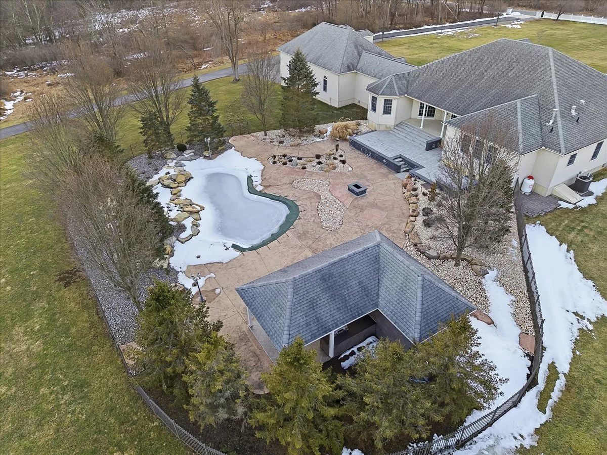 Aerial view of a backyard with a small frozen pond, rocks, trees, a patio, and a shed, with snow on the ground surrounding it.