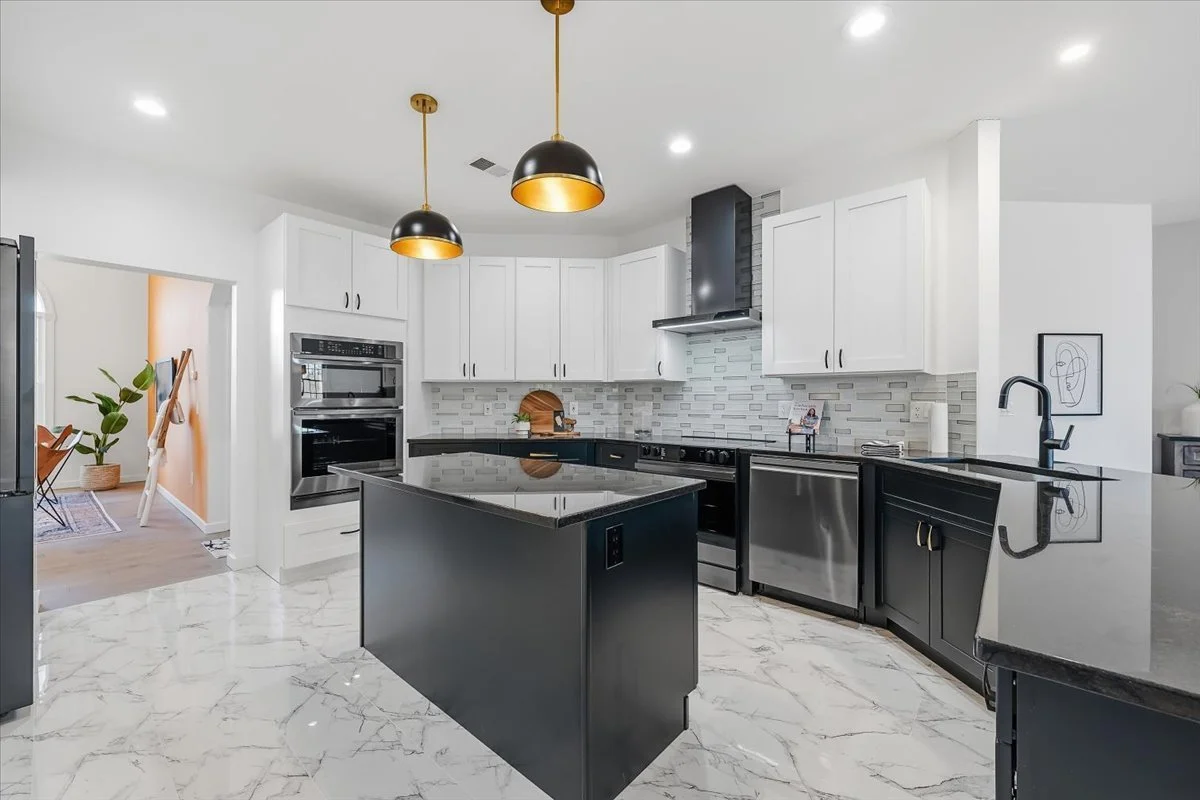 Modern kitchen with white upper cabinets, black lower cabinets, a black island with a marble top, stainless steel appliances, a black range hood, and pendant lights with black and gold accents.