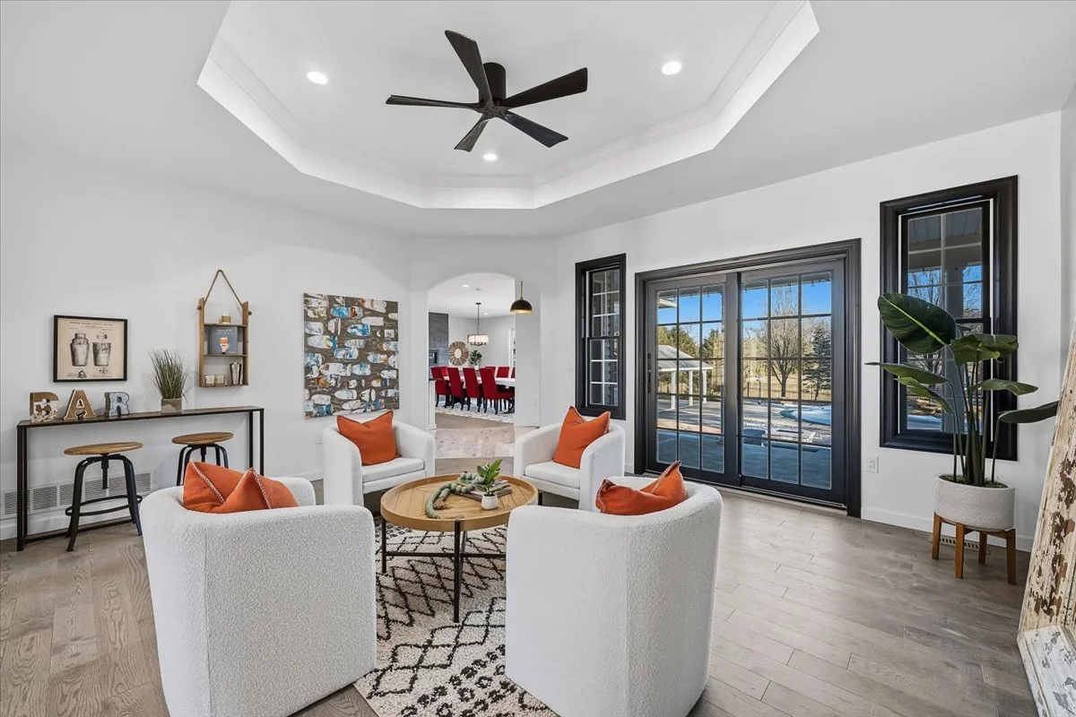 Bright living room with white walls, large black framed windows, and a sliding glass door. Features a circular ceiling with recessed lighting and a black ceiling fan. Contains four white armchairs with orange pillows around a round wooden coffee tabl