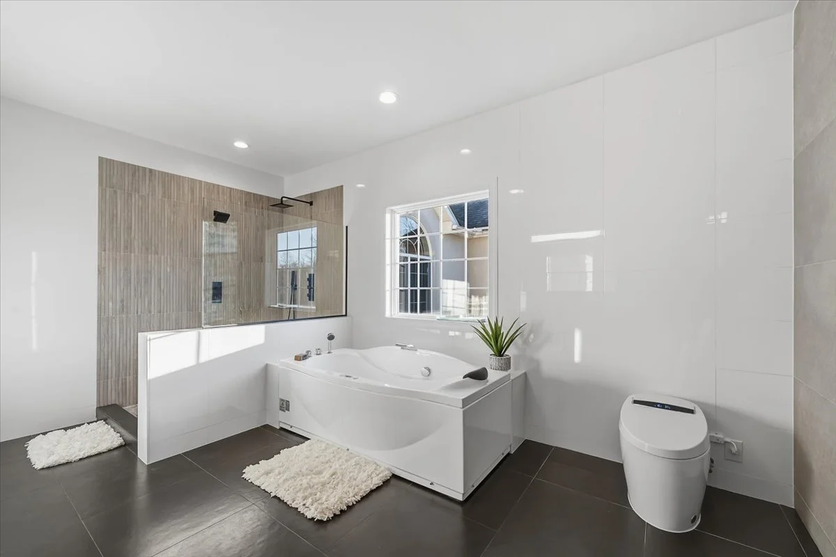 Modern bathroom with white walls, a large window, a corner bathtub, a walk-in shower with a glass partition, a small potted plant, dark tile flooring, and two white bath mats.