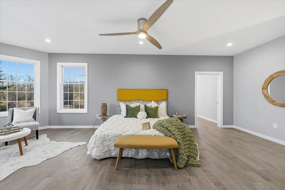 A modern bedroom with light gray walls, hardwood flooring, a large bed with a yellow headboard, white bedding, green and white pillows, a knitted green throw, and a yellow bench at the foot of the bed. There are two windows on the left, a white chair